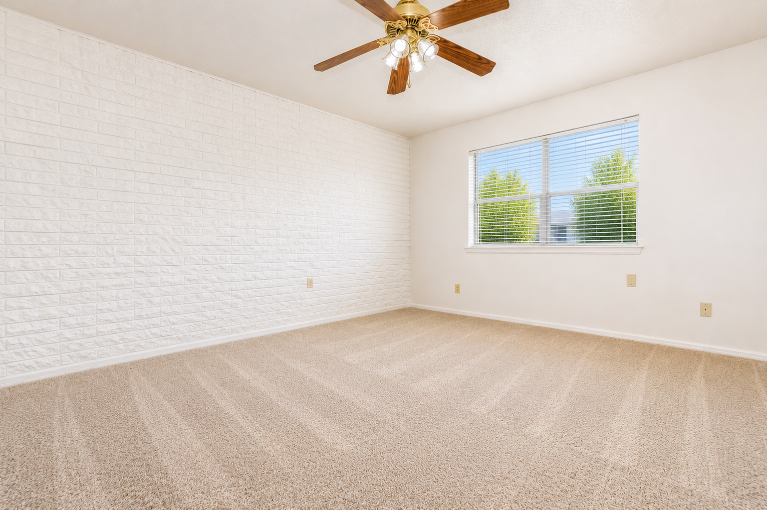 Empty room with a light-colored carpet and a white brick wall. A ceiling fan with light fixtures is visible, and there is a window with white blinds, showcasing green trees outside. The walls are painted white, creating a bright and spacious atmosphere.