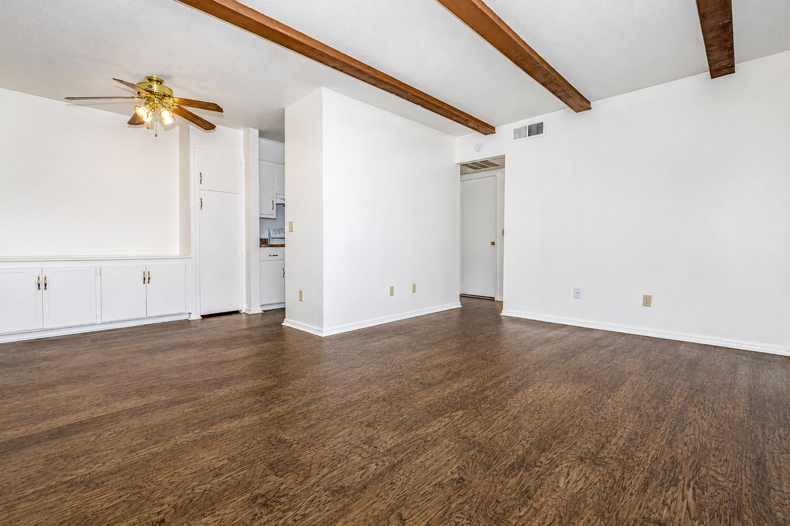 Spacious, empty living room with wooden flooring, white walls, and exposed ceiling beams. A ceiling fan hangs above, and there are built-in cabinets on one side. A doorway leads to another room, and the kitchen is visible in the background. Natural light fills the space, creating a bright and inviting atmosphere.