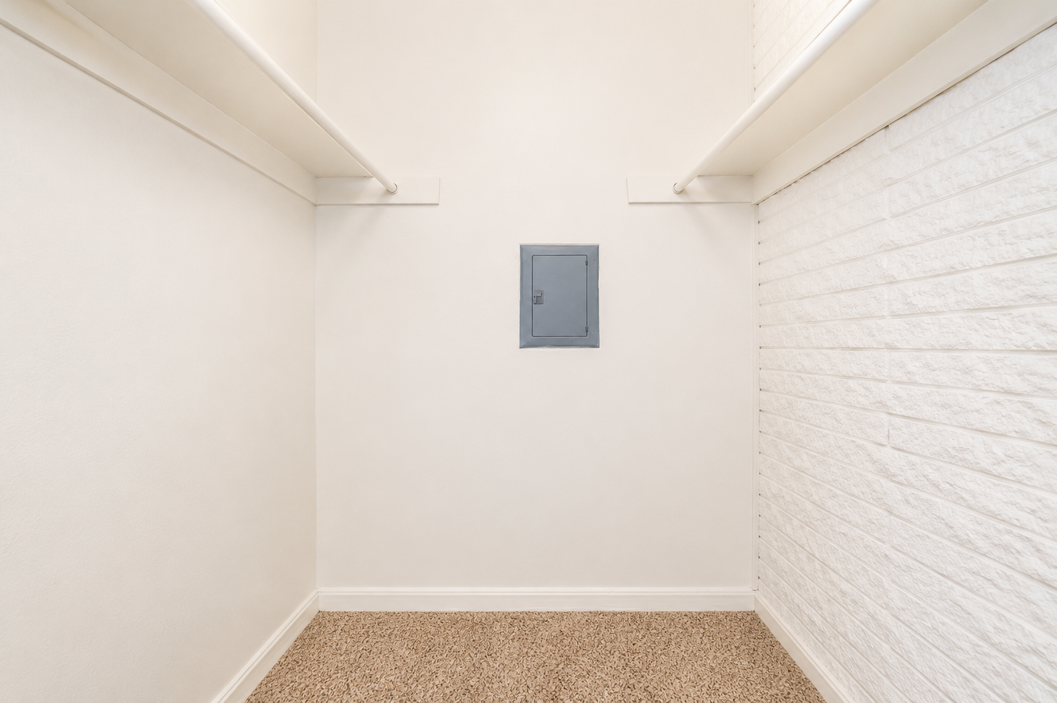 A minimalistic room with white walls and a light brown carpet. On the back wall, there is a small gray door or access panel. The space features two hanging rods on either side, emphasizing its emptiness and simplicity.