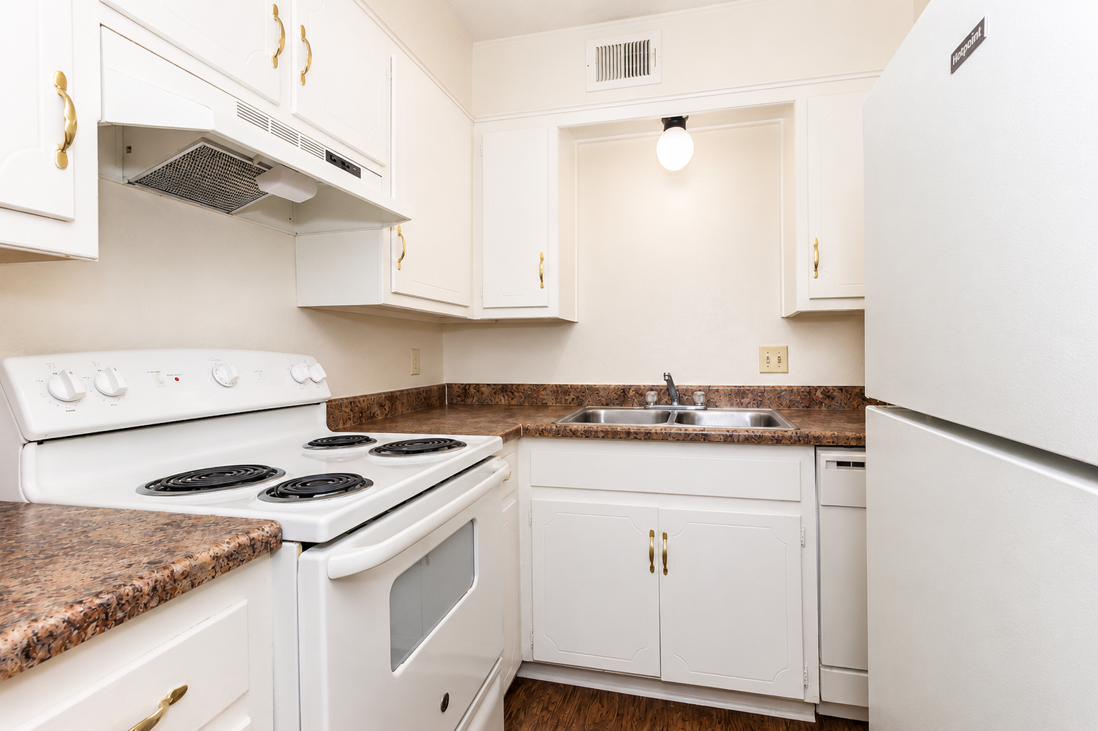 A bright and clean kitchen featuring white cabinetry, a white stove with four burners, and a matching refrigerator. The countertop is made of a dark textured material, and a double sink is situated under a light fixture. The kitchen has a modern and well-organized appearance.