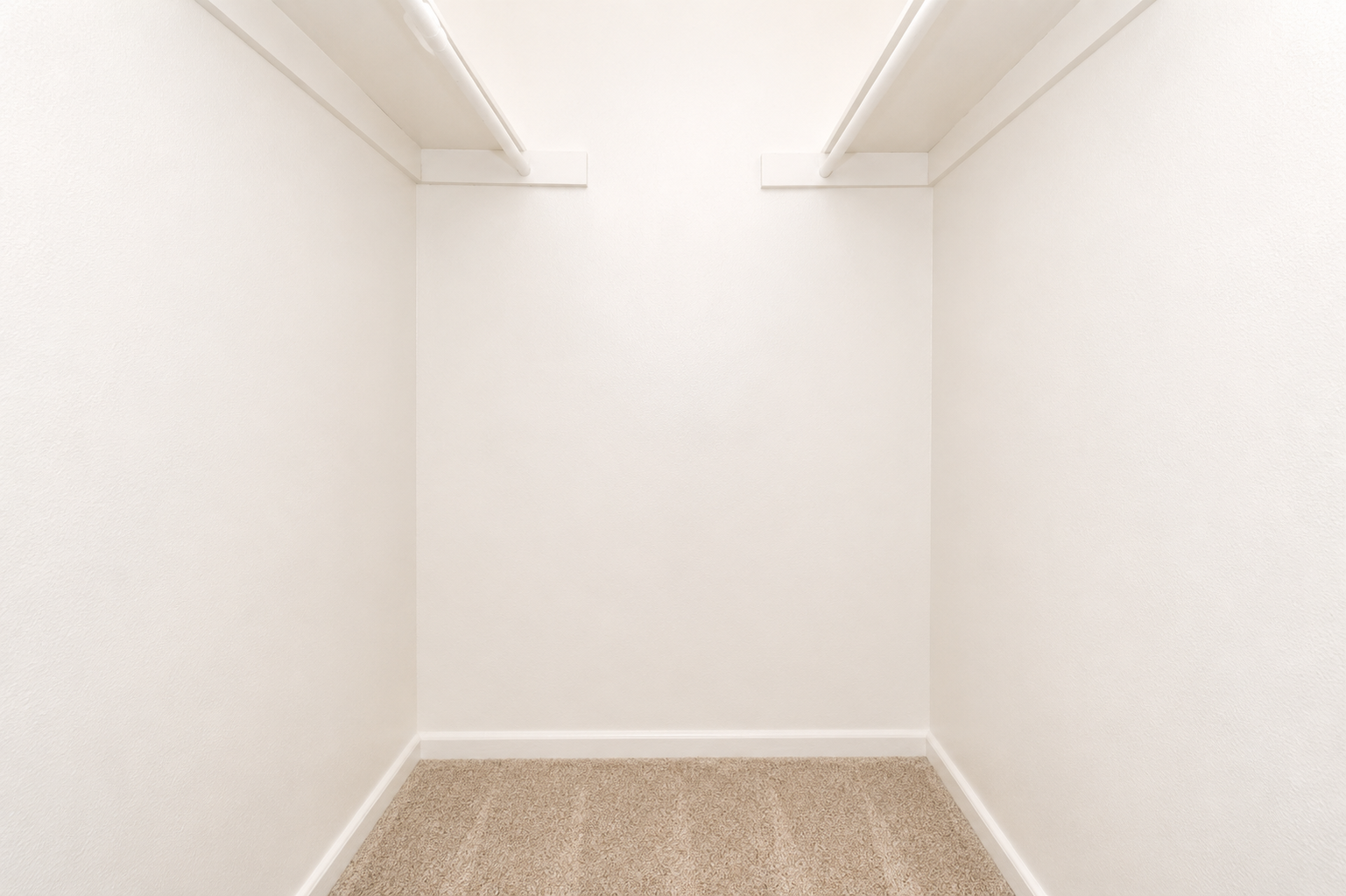 Empty interior of a closet with white walls and a tan carpeted floor. The space is well-lit, with no furnishings or items visible. Shelves are mounted on the upper wall, creating a clean and minimalistic look.