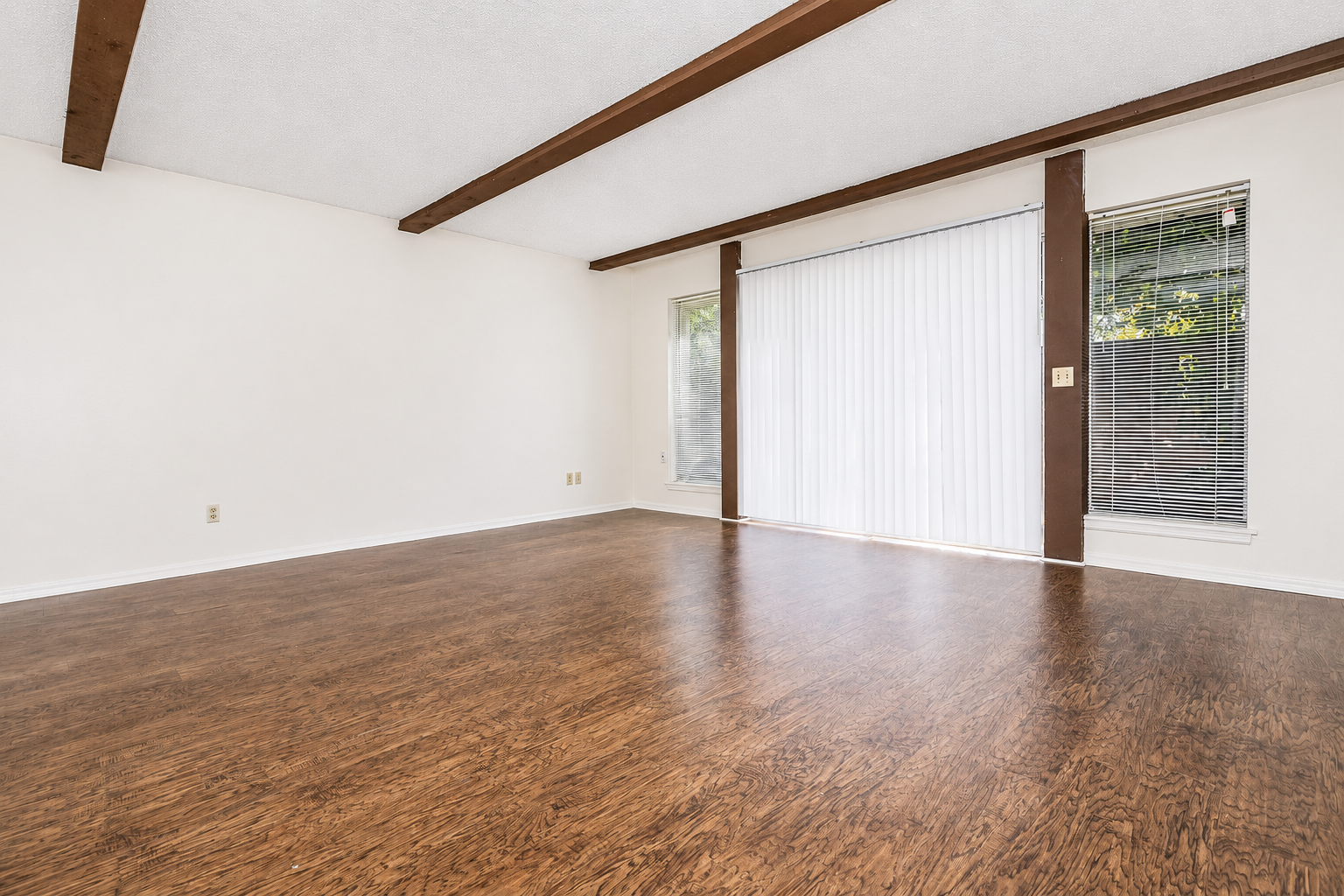 Spacious, empty living room featuring light-colored walls and wooden beams on the ceiling. The floor is a rich wood laminate. A large window with vertical blinds allows natural light, and there's a sliding door leading outside. The room is designed for flexibility in furnishing and decoration.