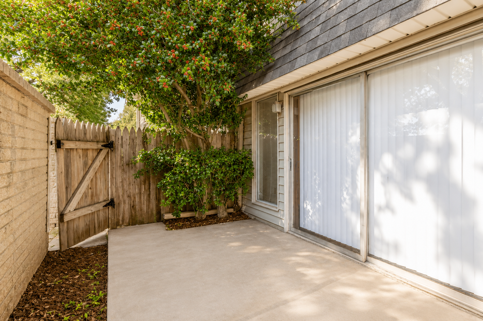A small outdoor patio area featuring a concrete surface, partially surrounded by a wooden fence. There is a bushy tree with green leaves on one side and a set of sliding glass doors leading into an interior space. Sunlight filters through, casting shadows on the patio.