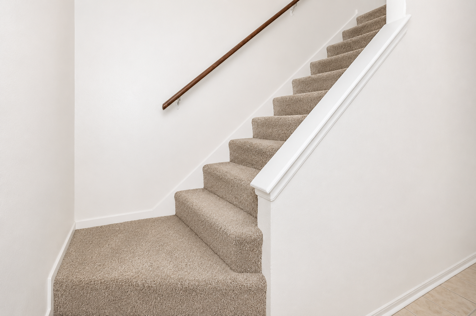 A carpeted staircase leading upward, featuring a wooden handrail on the right. The walls are plain and white, and the steps are covered in a light beige carpet, providing a clean and simple aesthetic. The flooring at the bottom is a light-colored tile.