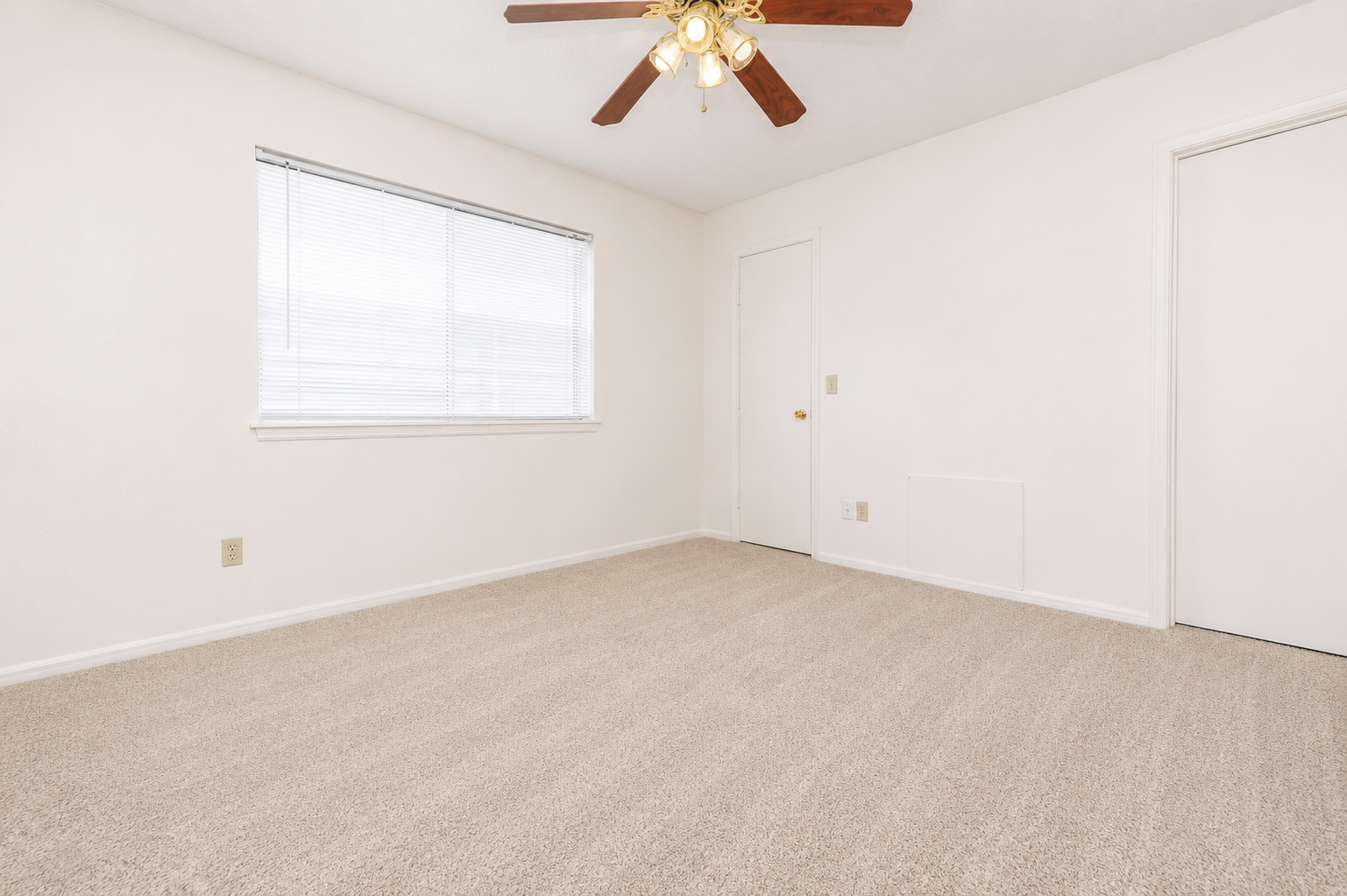 Empty room with light beige carpet and white walls. A ceiling fan with wooden blades is mounted in the center. One window with blinds allows natural light, and a closed door is visible on the right side. The space appears clean and unfurnished, offering a bright and open feel.