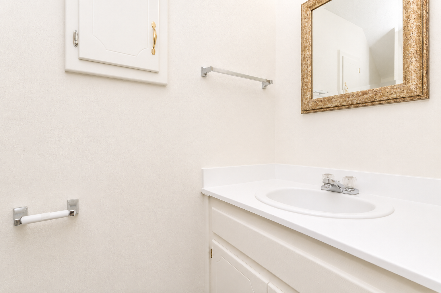 A clean, bright bathroom featuring a white countertop with an oval sink, a decorative mirror with a gold frame, and a wall-mounted towel rack. The walls are a light neutral color, and there is a white cabinet above the sink. A toilet paper holder is visible on the side.