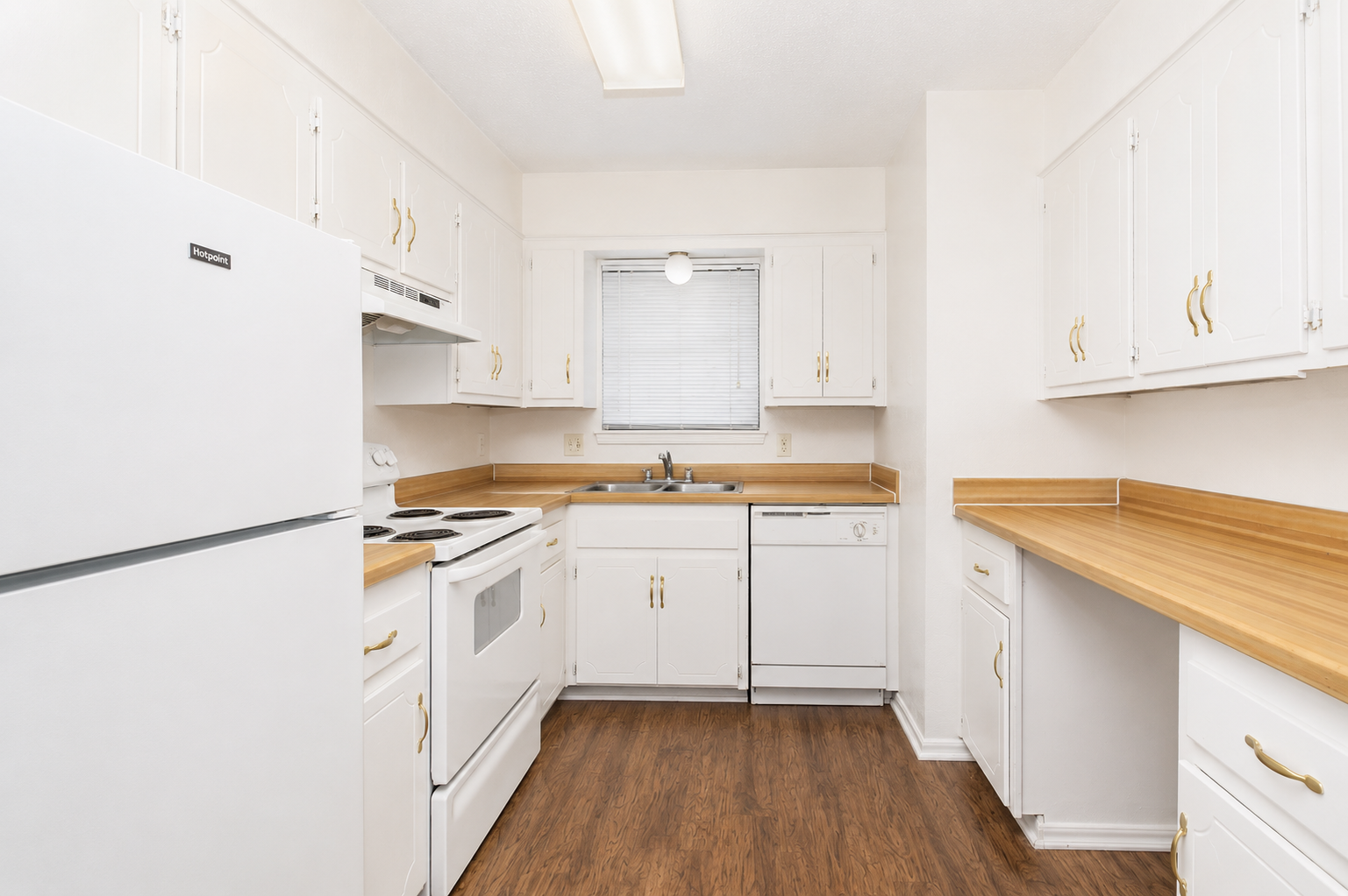 Bright and clean kitchen featuring white cabinetry with gold handles, a matching white refrigerator, stove, and dishwasher. The countertops are light wood, and there's a window above the sink providing natural light. The flooring is a dark wood laminate, giving a modern and cozy appearance.