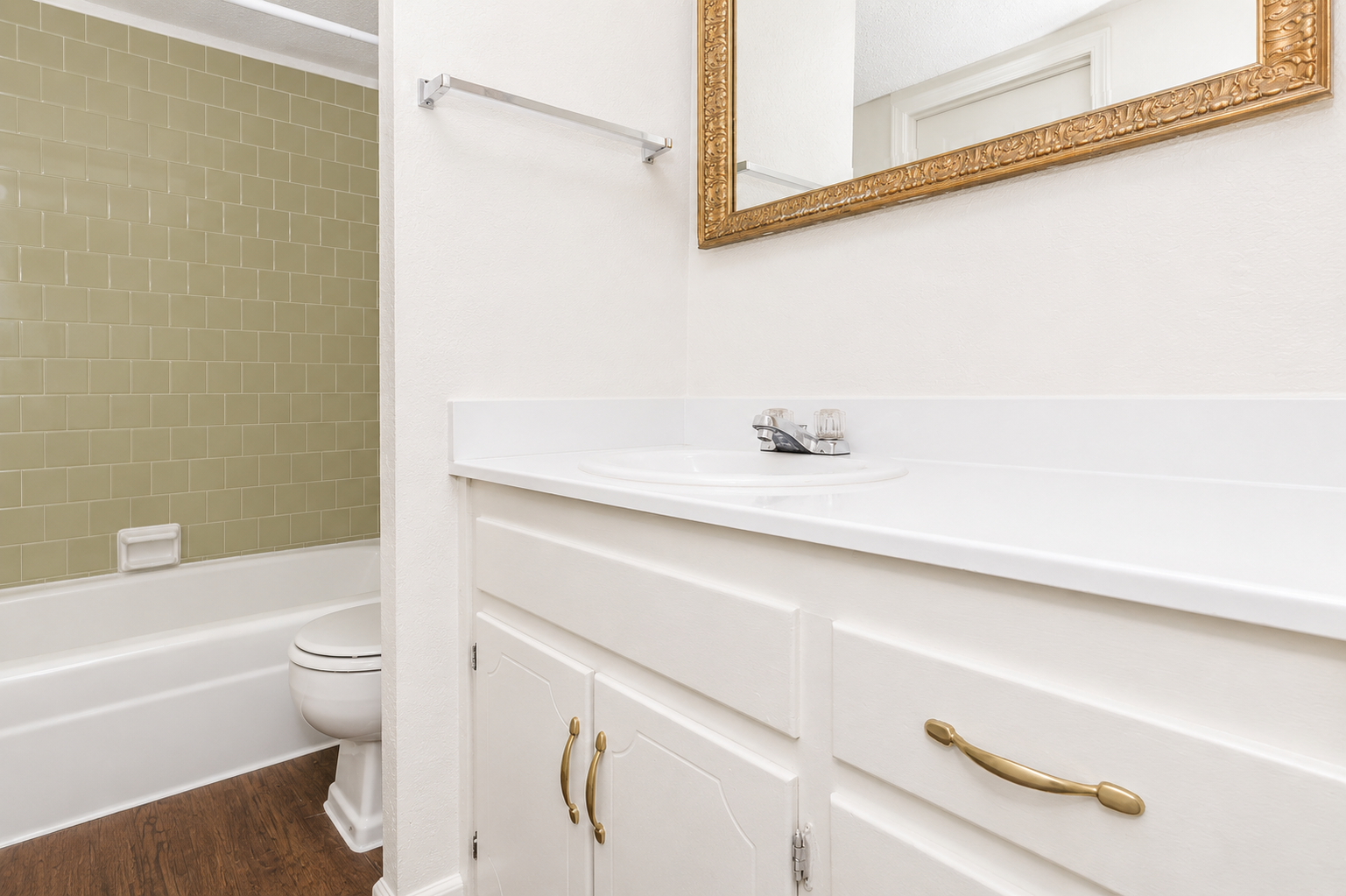A clean bathroom featuring a white sink with a faucet, a wall-mounted mirror in a gold frame, and light-colored cabinetry. In the background, there is a bathtub with green tile surround and a white toilet. The flooring is a dark wood finish, creating a contrast with the bright bathroom elements.