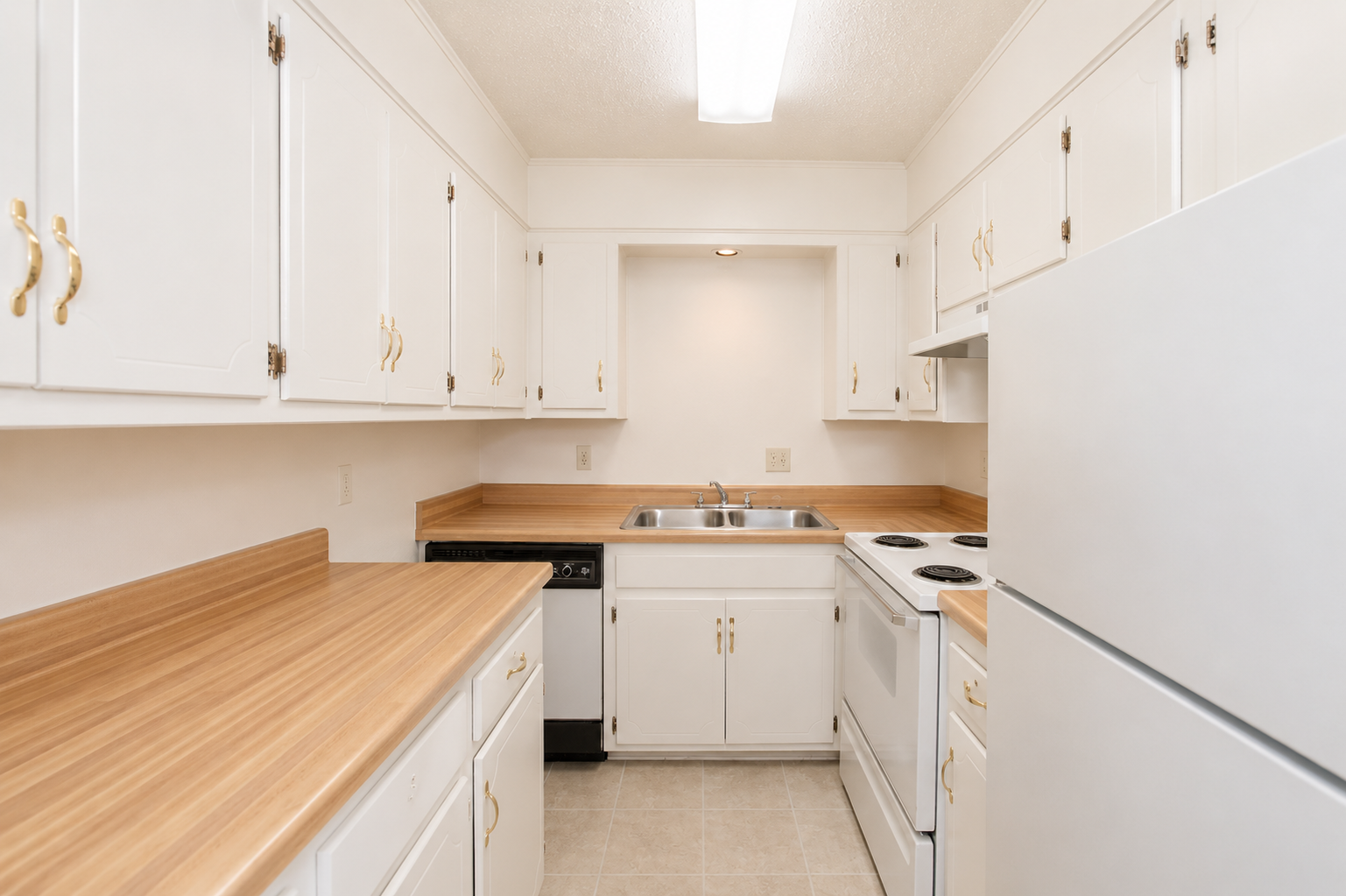 A small, modern kitchen with white cabinets and beige countertops. The kitchen features a sink, stove, and refrigerator. There are empty spaces for appliances, and the walls are painted a light color, creating a bright and clean atmosphere. The flooring is light-colored tile.