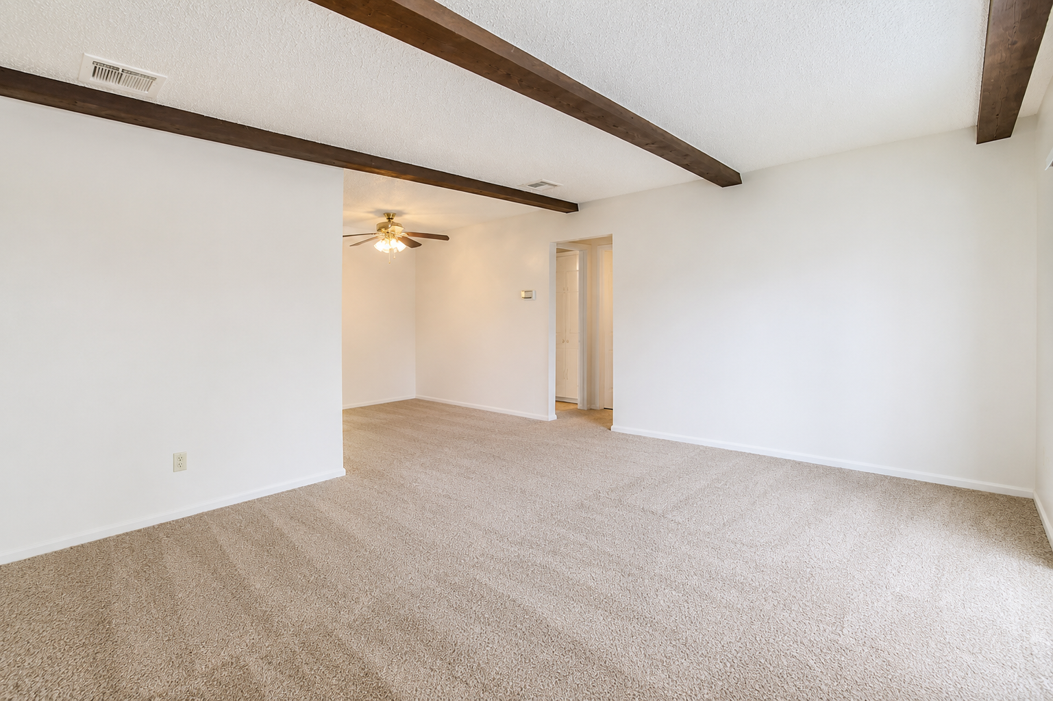 A spacious, unfurnished room with light-colored walls and a textured ceiling. The floor is covered in beige carpet. A ceiling fan is mounted in one corner, and there are exposed wooden beams along the ceiling. A doorway at the rear leads to another room or hallway. Natural light is coming in from the left side.
