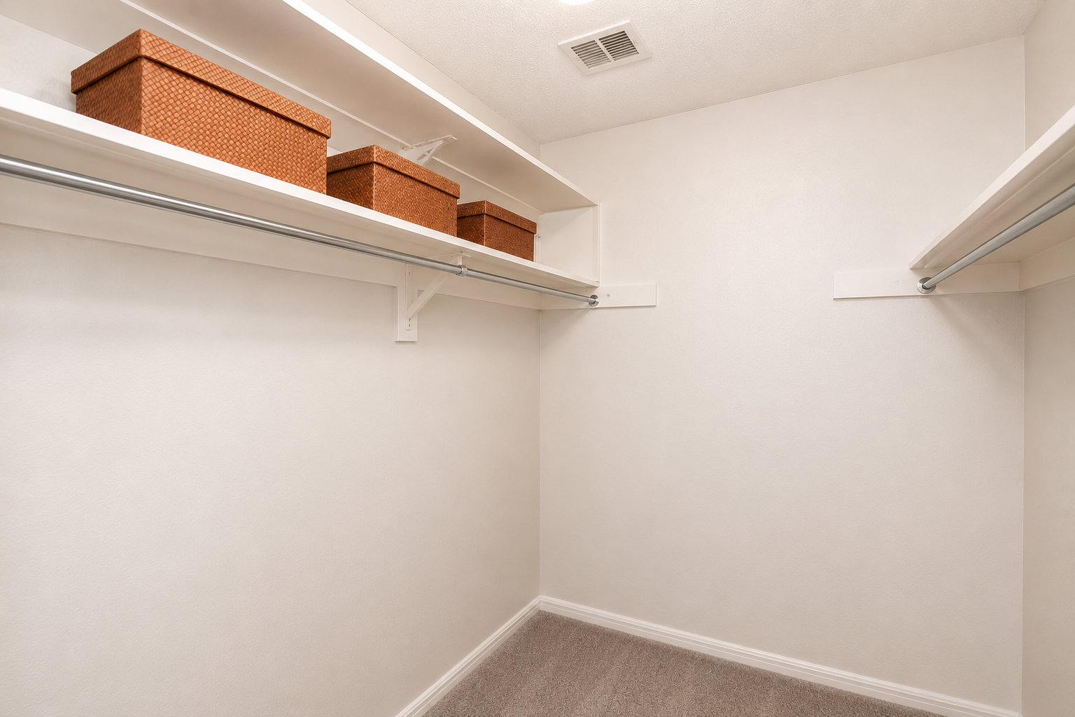 A clean, empty closet with light-colored walls and a plush carpet. There are two metal hanging rods on either side, and several decorative storage boxes arranged neatly on the upper shelf. The overall atmosphere is organized and spacious.