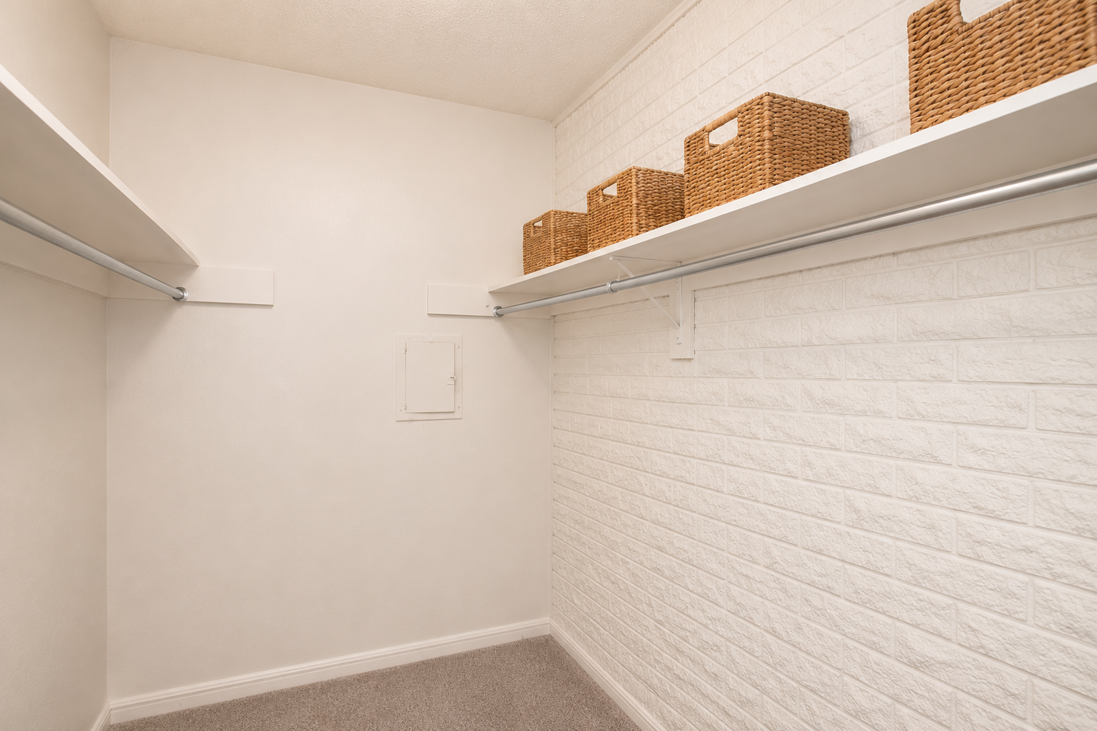 A clean and empty closet featuring white walls and a textured accent wall. Two shelves are mounted on the walls, holding a few wicker storage baskets. The floor is carpeted, and there is a small access panel on the wall. The space is well-lit and organized.