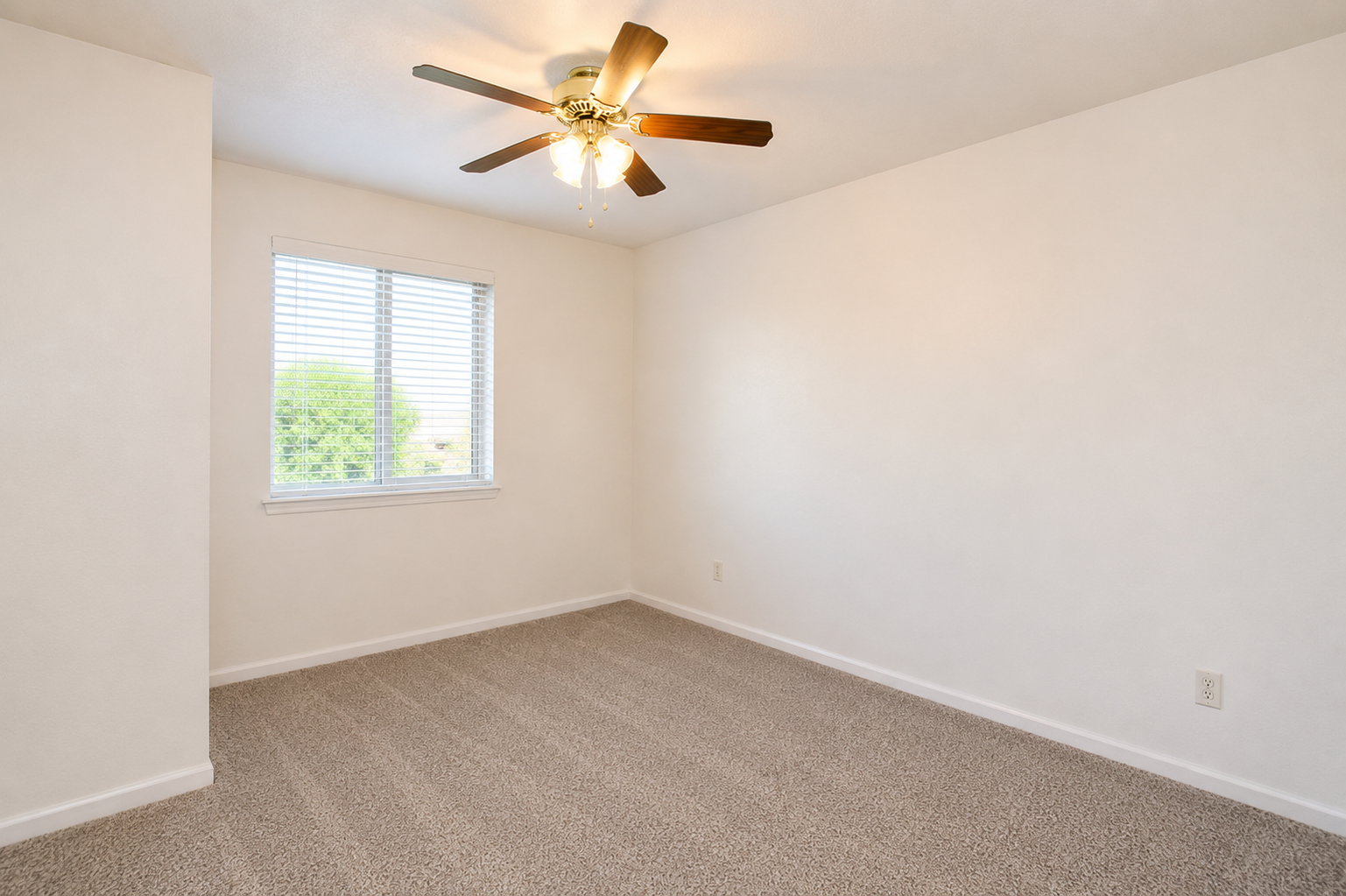A well-lit empty room with beige carpet flooring and white walls. It features a ceiling fan with light and a window with blinds, offering a view of greenery outside. The space is open and ready for furnishing.