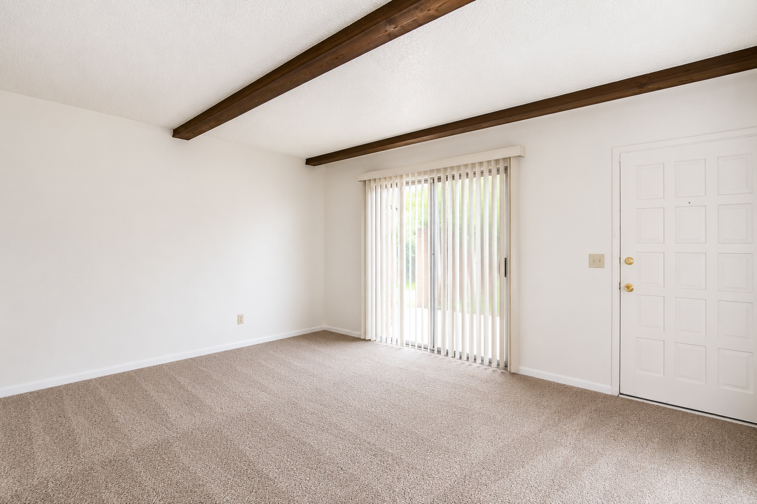A spacious room with light beige carpet and white walls. The ceiling features wooden beams, and there are sliding glass doors with vertical blinds leading to an outdoor area. A plain white door is visible on the right side of the image, creating a bright, inviting atmosphere.