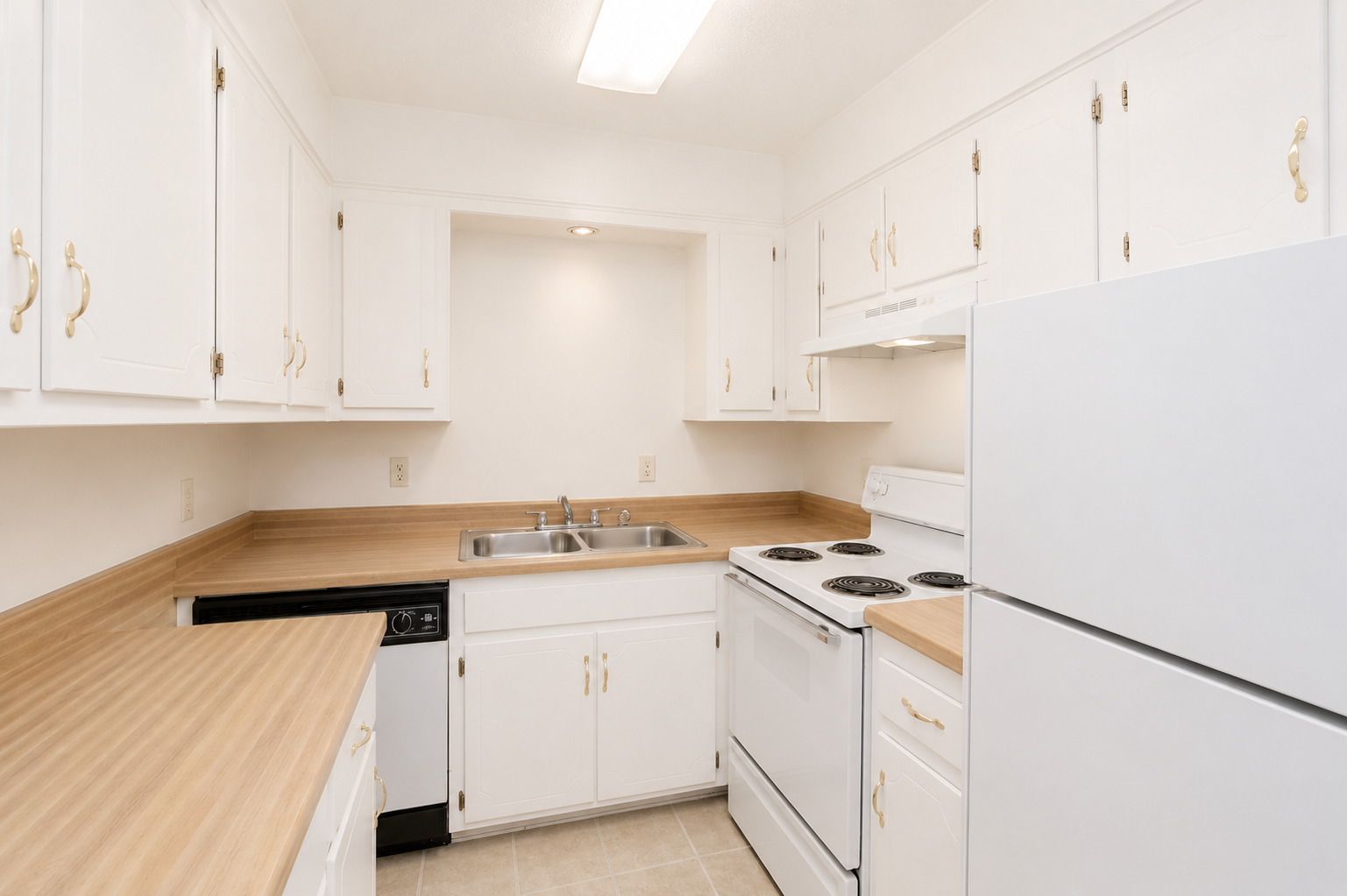A small, modern kitchen featuring white cabinetry, a beige countertop, stainless steel sink, and a white refrigerator. The kitchen includes a stove with an oven, a dishwasher, and a light fixture above the workspace. The floor is light-colored tile, creating a bright and clean atmosphere.