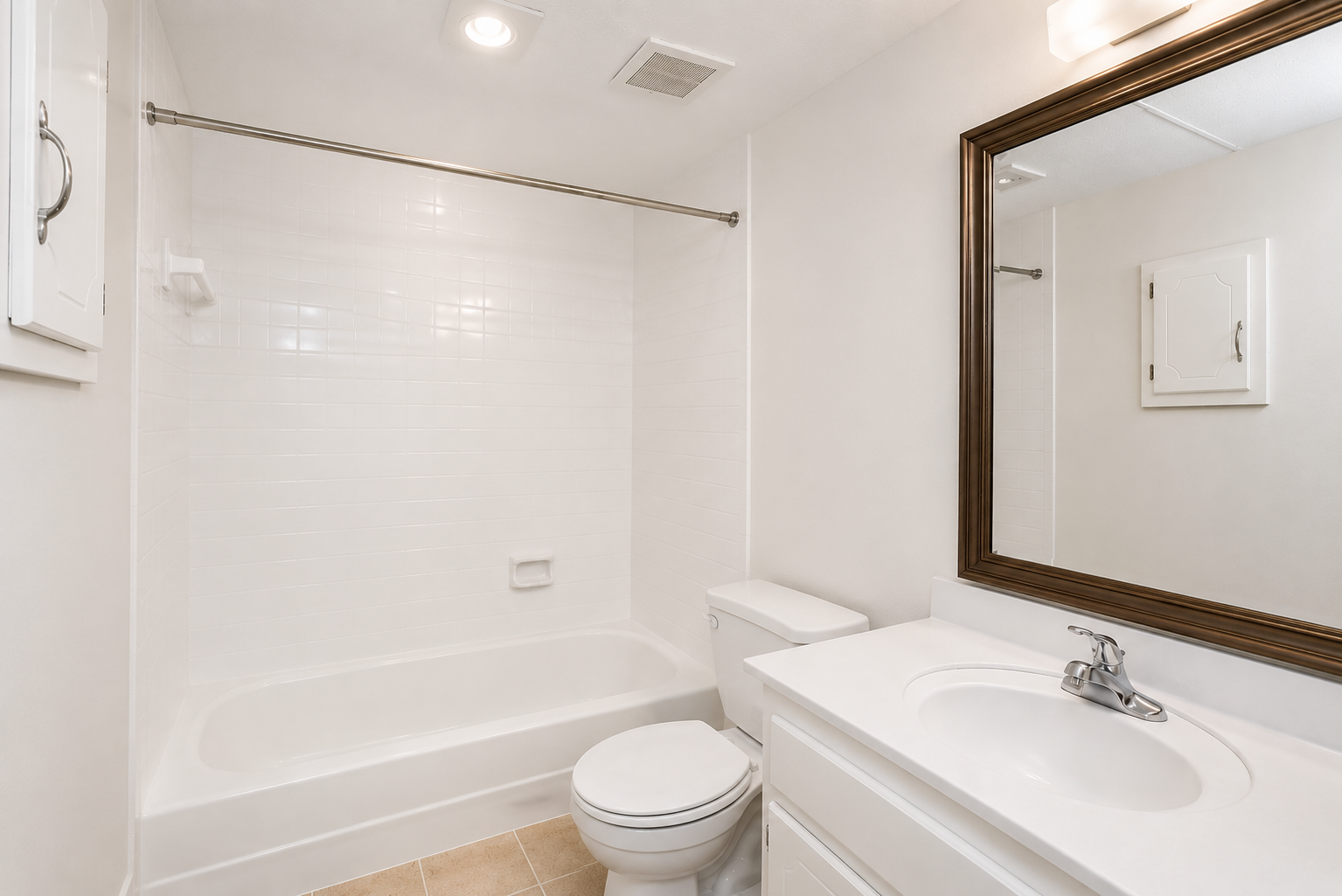 A clean and well-lit bathroom featuring a white bathtub with a shower, a modern toilet, and a single sink vanity with a mirror above. The walls are tiled in white, and there is a light fixture on the ceiling, creating a bright and inviting atmosphere.