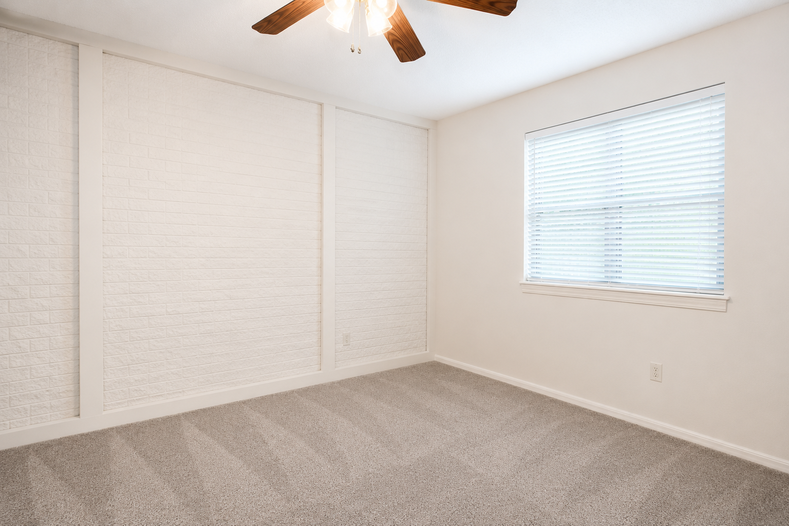 A bright and empty room featuring light-colored walls, a white brick accent wall, and a ceiling fan. The floor is covered with plush gray carpet, and there is a window with blinds allowing natural light to enter the space.