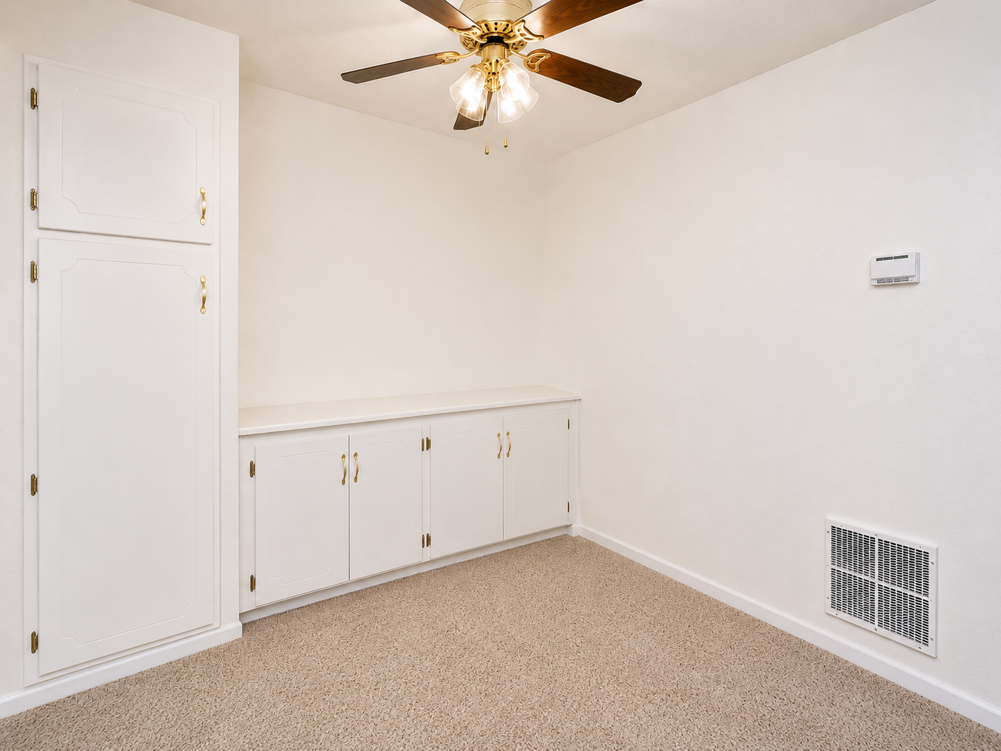 A light-colored room with beige carpet, featuring a ceiling fan with light fixtures. On one wall, there are white cabinets and a small shelf. An air vent is visible on the floor, and a thermostat is mounted on the adjacent wall. The room has a minimalistic and spacious appearance.