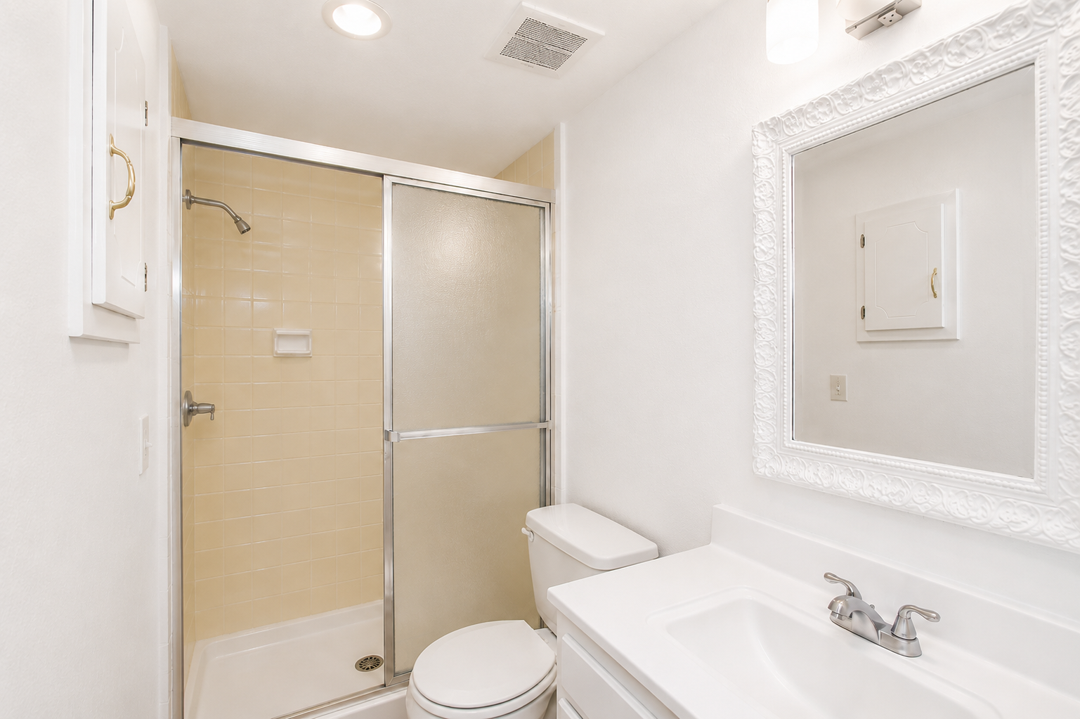 A small, tidy bathroom featuring a shower stall with a glass door, a white toilet, and a sink with a mirrored cabinet above. The walls are painted a light color, and there is overhead lighting. The floor is not visible.