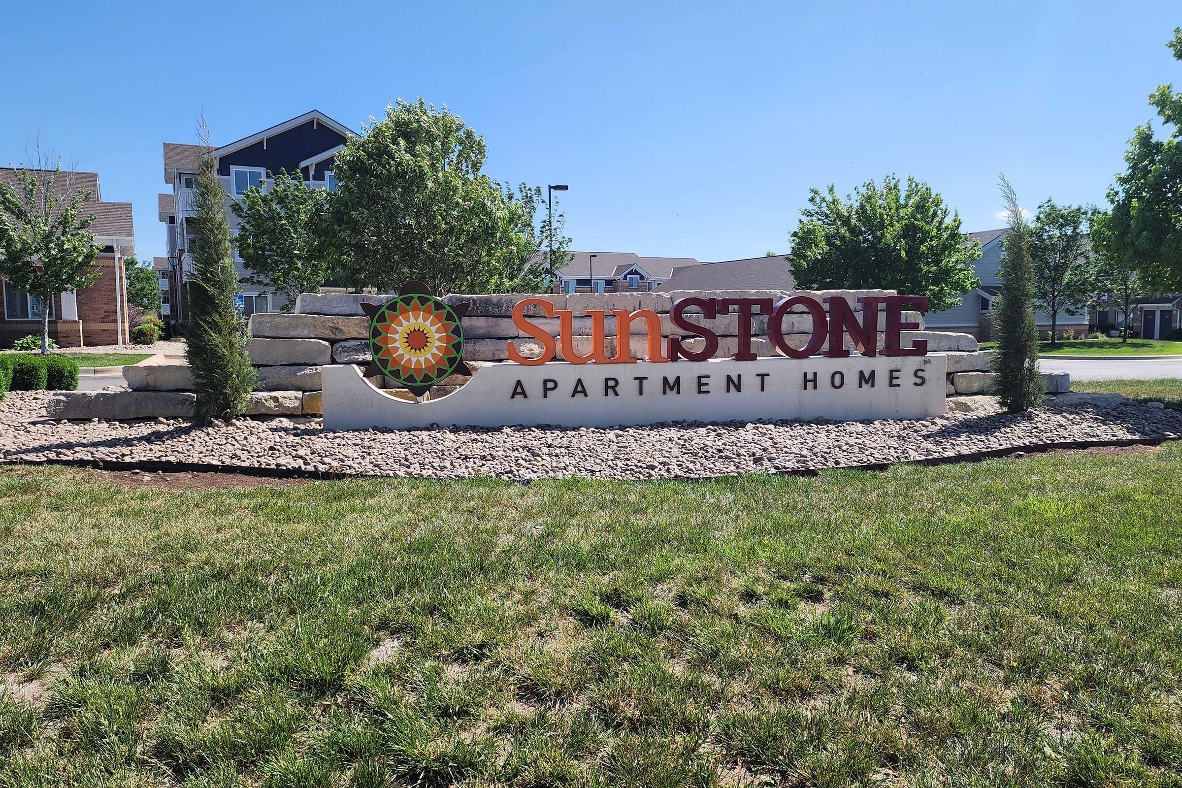 Sign for Sunstone Apartment Homes featuring a sun logo and surrounded by landscaping, including trees and decorative stones. Visible blue sky and a grassy area in the foreground.