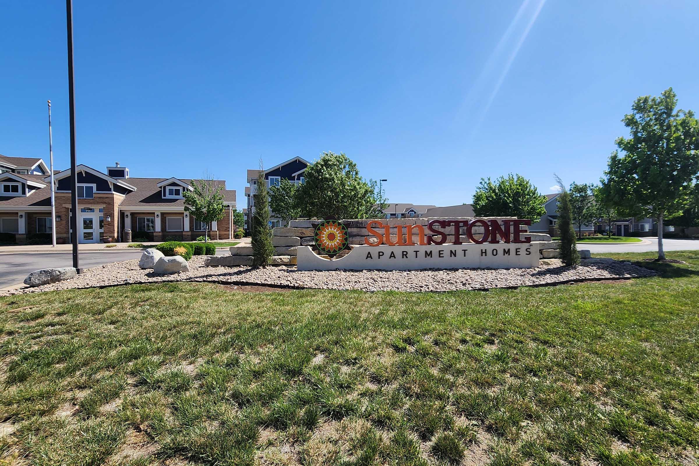 Sign for Sunstone Apartment Homes, featuring the name prominently displayed with a decorative sun emblem above it. Surrounding the sign, there are landscaped areas with shrubs and small rocks, set against a clear blue sky and a residential apartment complex in the background.