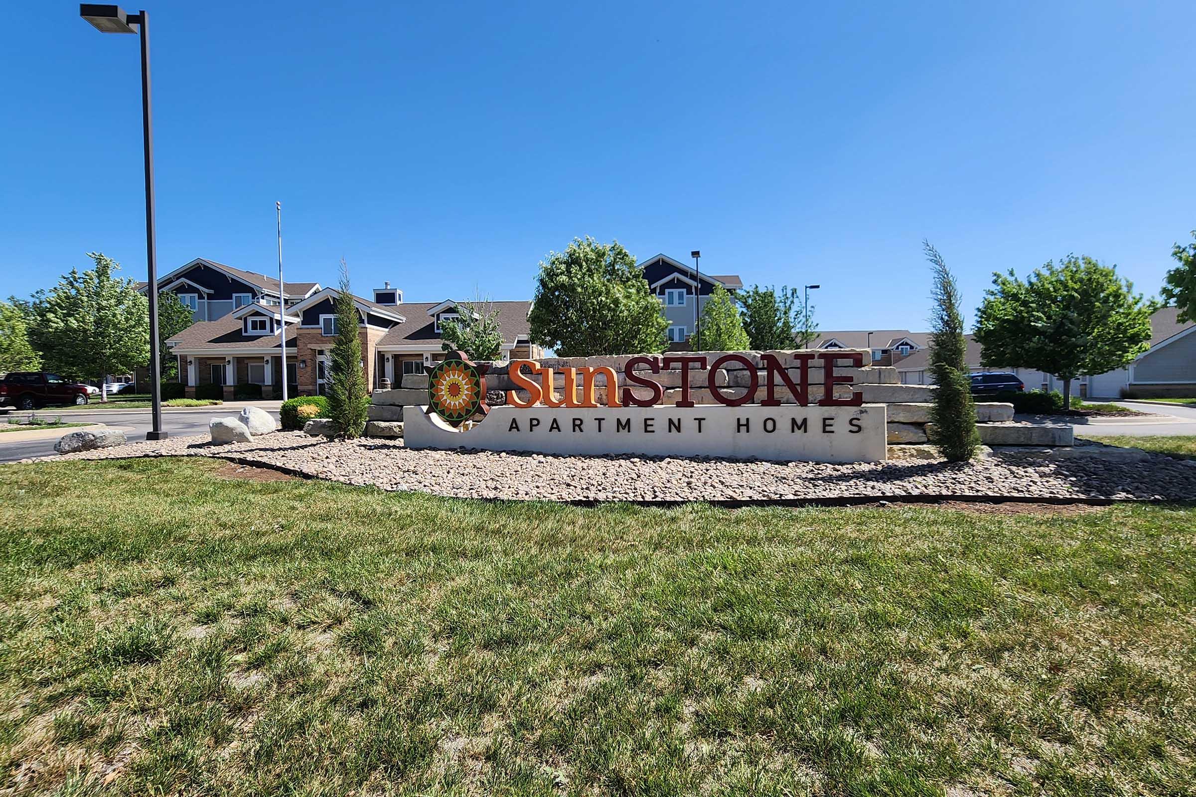 A sign for "Sunstone Apartment Homes" with a decorative sun logo, set against a clear blue sky. The landscaped area features grass, shrubs, and decorative rocks in front of a well-maintained apartment building visible in the background.