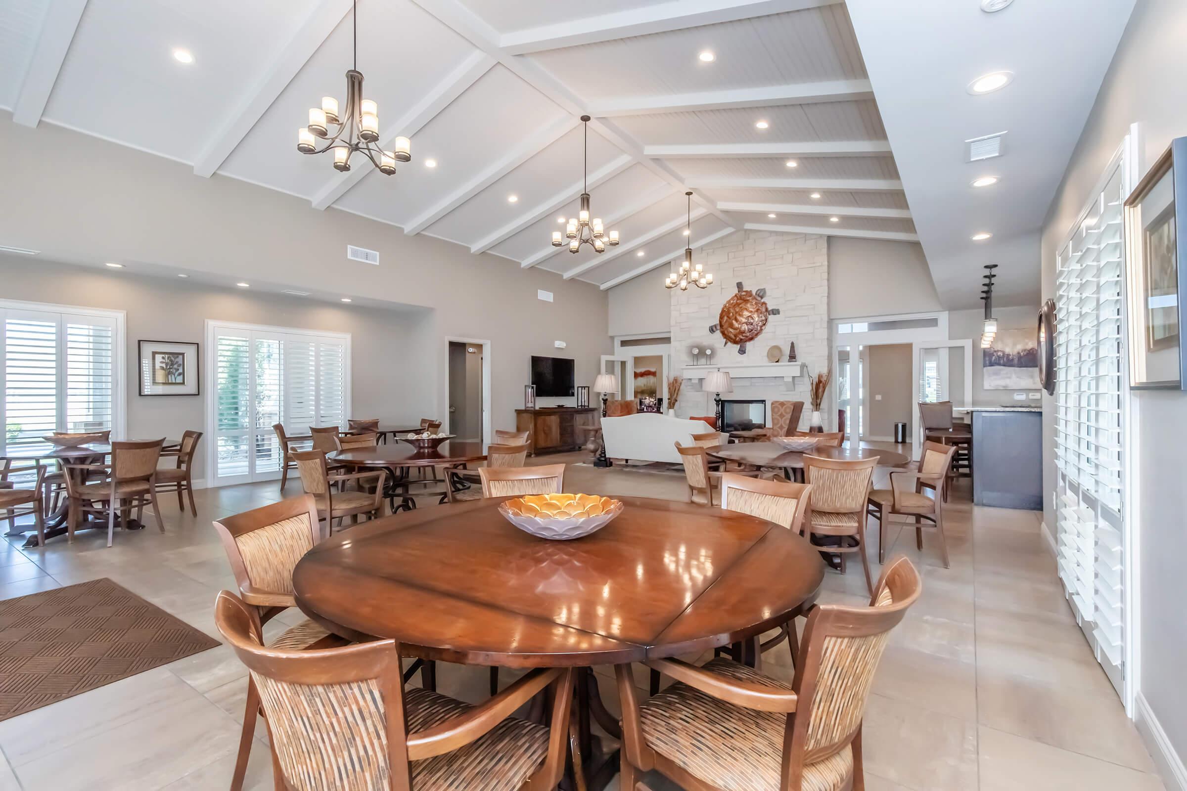 A spacious and elegantly designed common area featuring wooden tables and chairs, decorative light fixtures, a cozy seating arrangement by a fireplace, and large windows with shutters allowing natural light. The room has a neutral color palette and clean architectural lines.