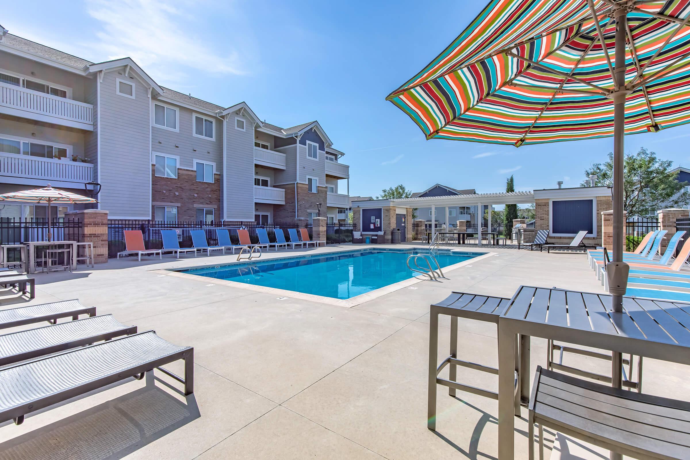A sunny outdoor pool area with lounge chairs and colorful umbrellas. Surrounding the pool are modern apartment buildings, creating a relaxing atmosphere for residents. The clear blue sky adds to the cheerful ambiance of the setting.