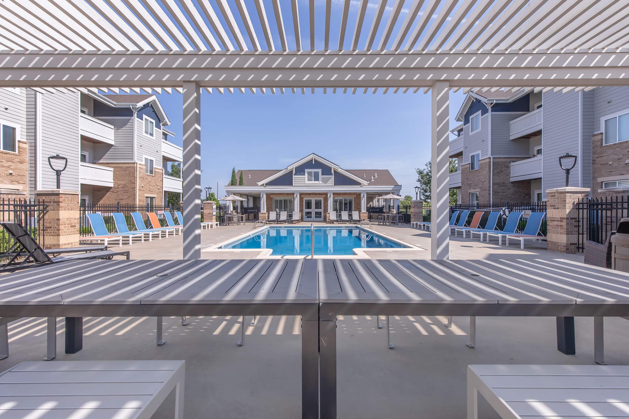 View of a modern outdoor swimming pool surrounded by residential buildings. Sun loungers are arranged around the pool area, and a shaded seating area with a slatted pergola is in the foreground. The sky is clear, creating a bright and inviting atmosphere for relaxation.