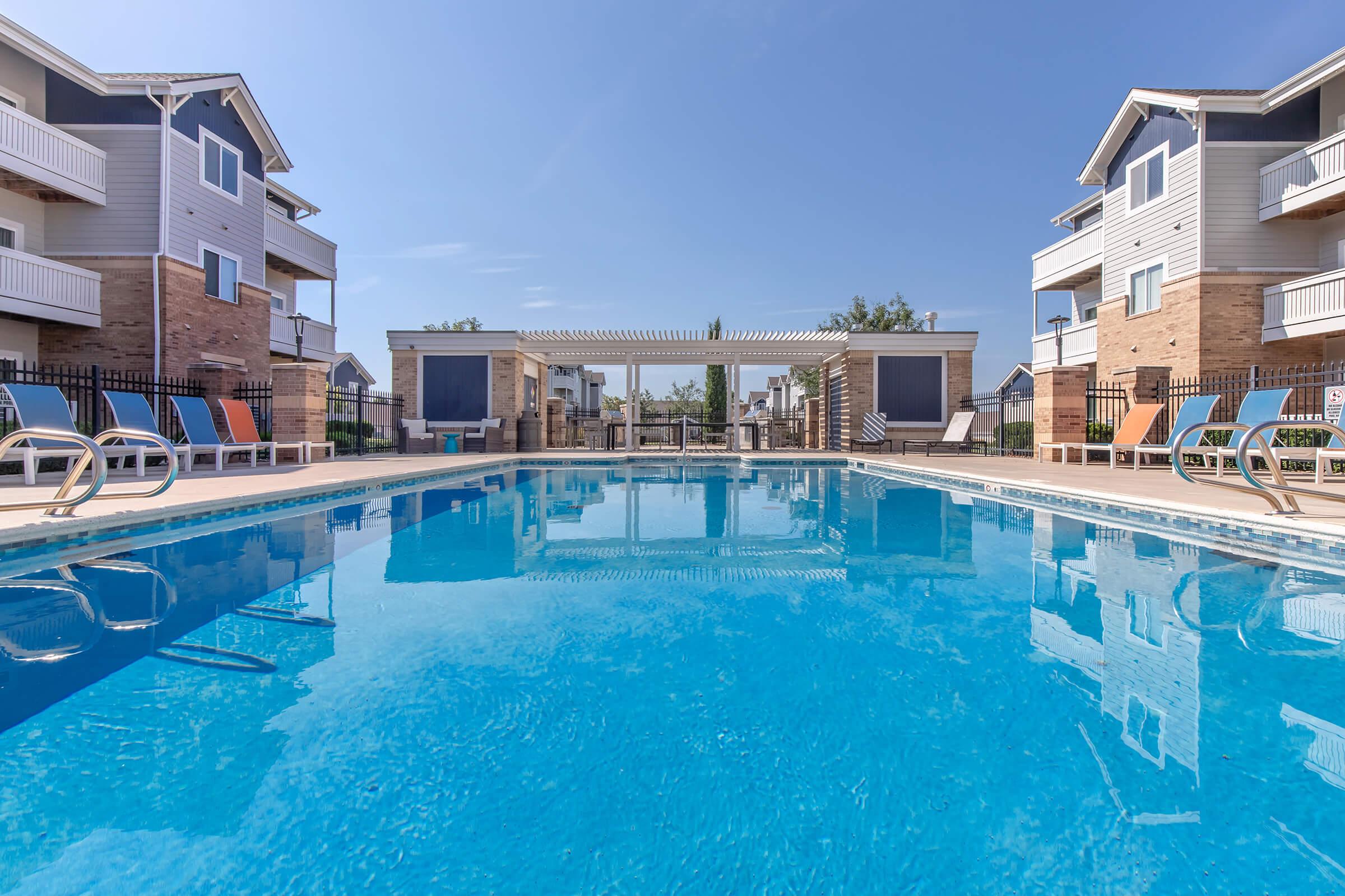 A clear blue swimming pool surrounded by lounge chairs, with two multi-story apartment buildings in the background. The sky is sunny and blue, and there are shaded areas provided by pergolas near the pool. The scene conveys a tranquil and inviting atmosphere for relaxation and leisure.