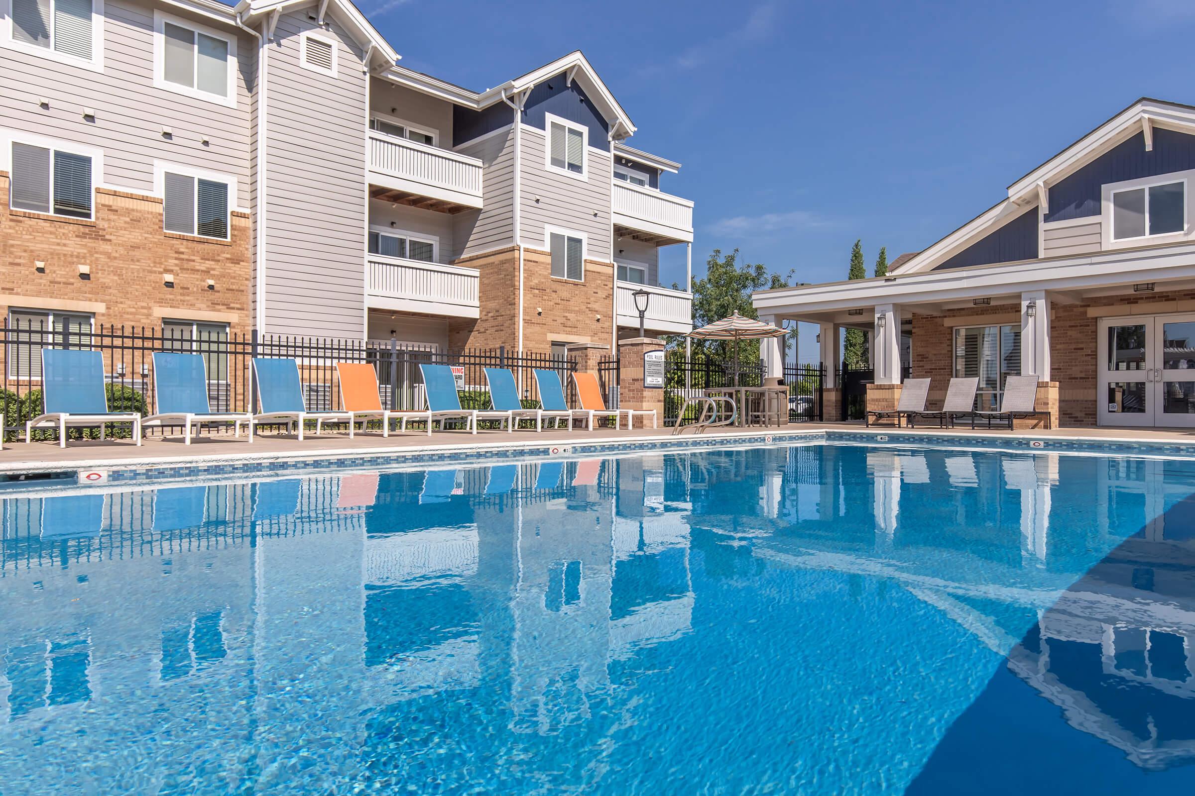 A clear blue swimming pool in front of a modern apartment complex. Lounge chairs in various colors line the poolside, with a shaded seating area and umbrellas. Bright blue sky reflected in the water creates a cheerful atmosphere.