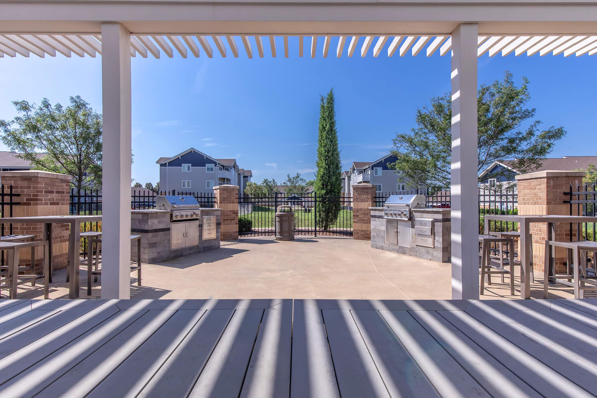 Outdoor patio area featuring a shaded structure, equipped with grilling stations and bar stools. The space is surrounded by greenery and a gated entrance, providing a welcoming environment for gatherings. Clear blue skies are visible in the background.