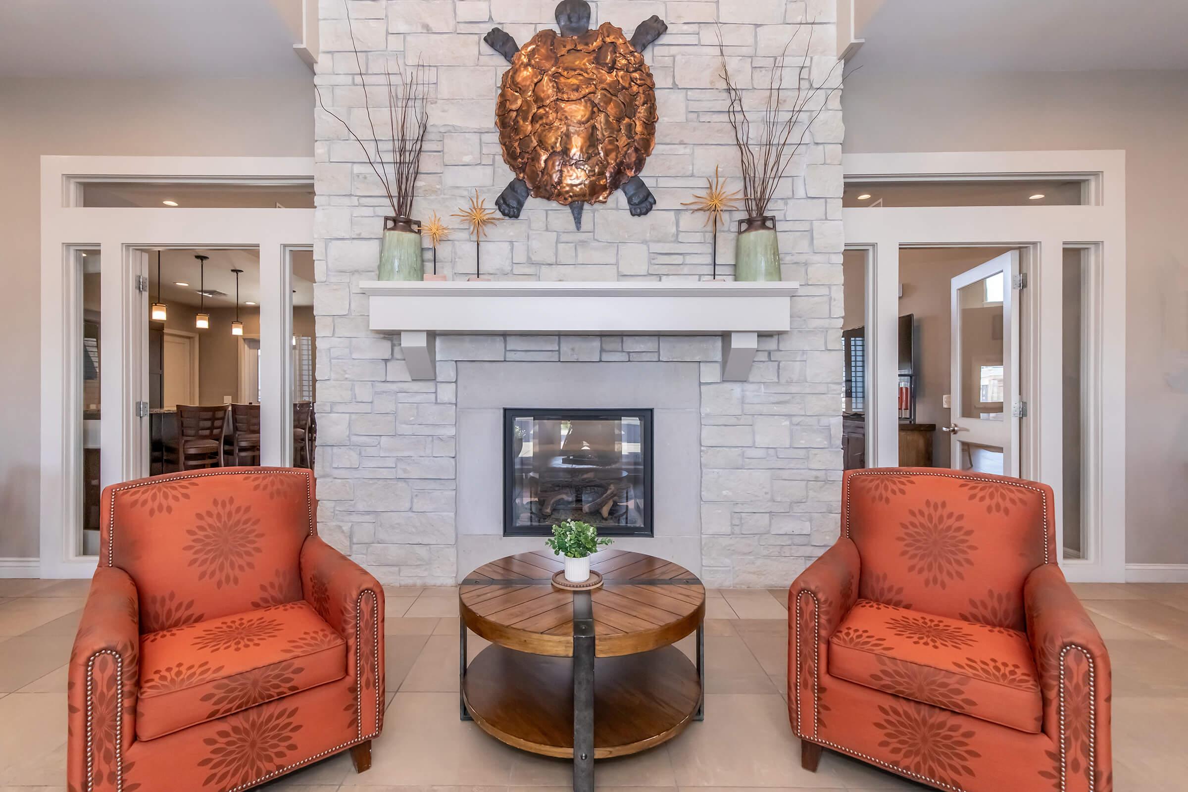 Two orange upholstered chairs with decorative patterns are positioned around a circular wooden coffee table. A stone fireplace has a turtle sculpture mounted above it, and there are decorative plants in vases on the mantel. The setting appears warm and inviting, with a modern aesthetic.