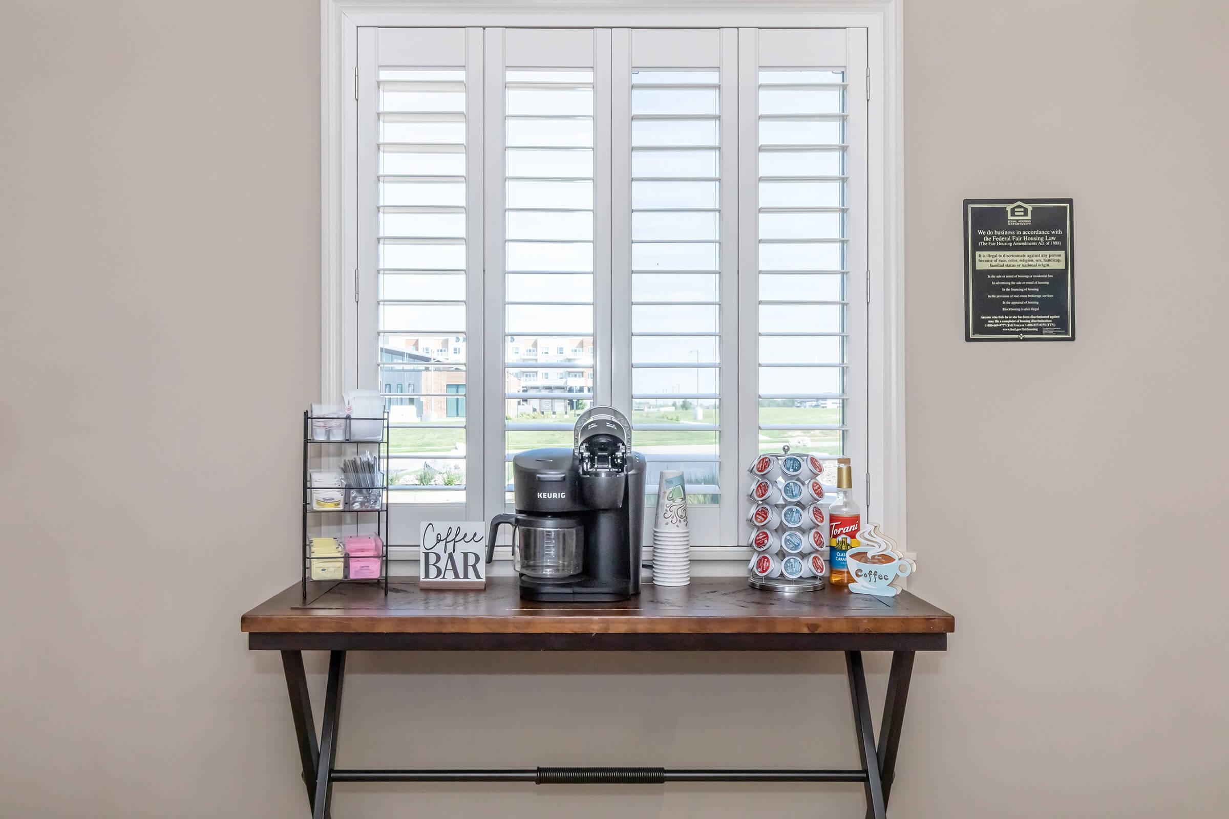 A coffee bar setup on a wooden table featuring a coffee machine, cups, and various coffee supplies. It is positioned in front of a window with plantation shutters, providing a view of the outdoors. A plaque is mounted on the wall nearby, and decorative elements enhance the cozy atmosphere.