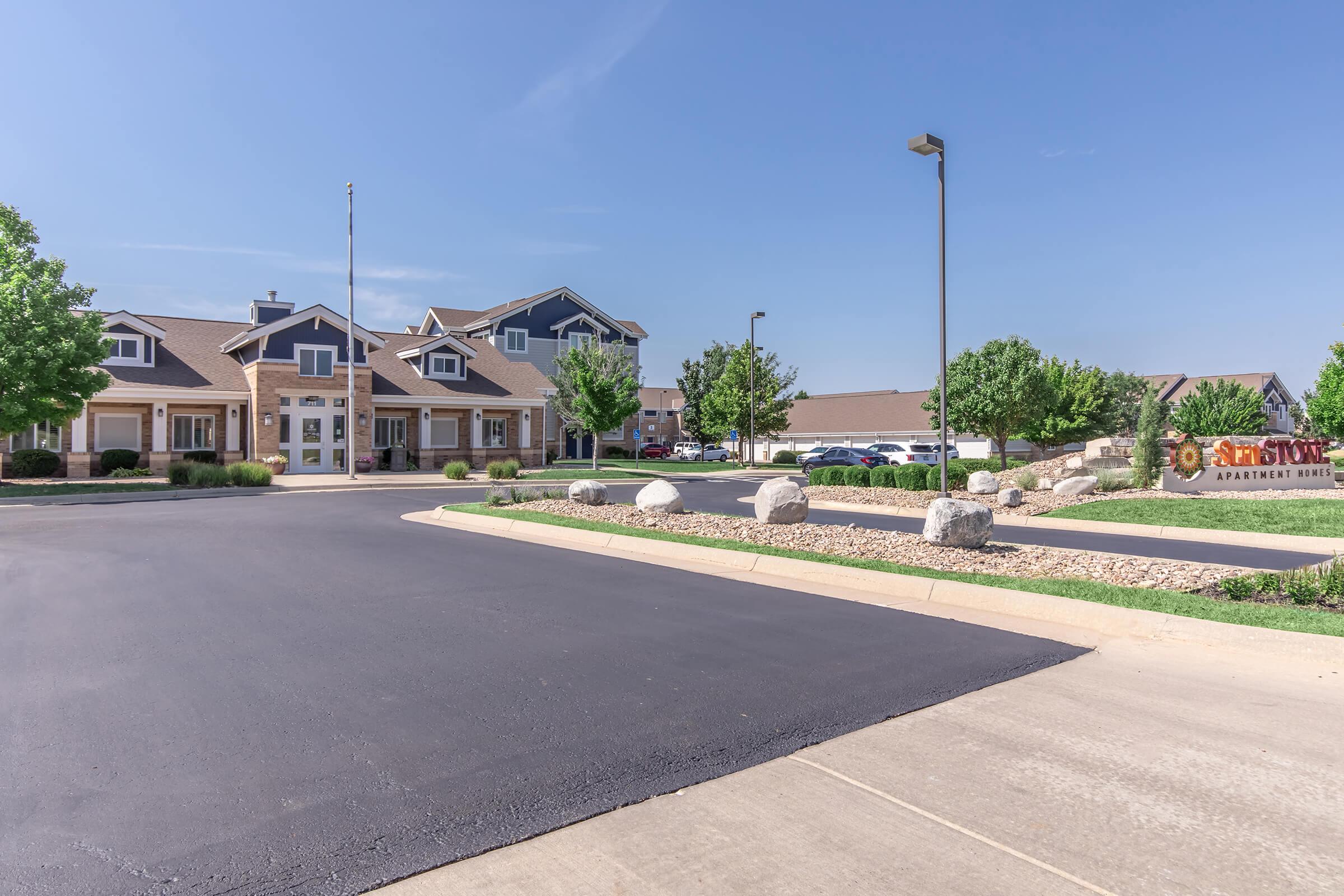 Apartment complex entrance with a well-maintained parking lot and landscaped greenery. The building features a mix of modern and traditional architectural styles, with a flagpole and several large boulders in the foreground. Clear blue sky overhead adds to the welcoming atmosphere.
