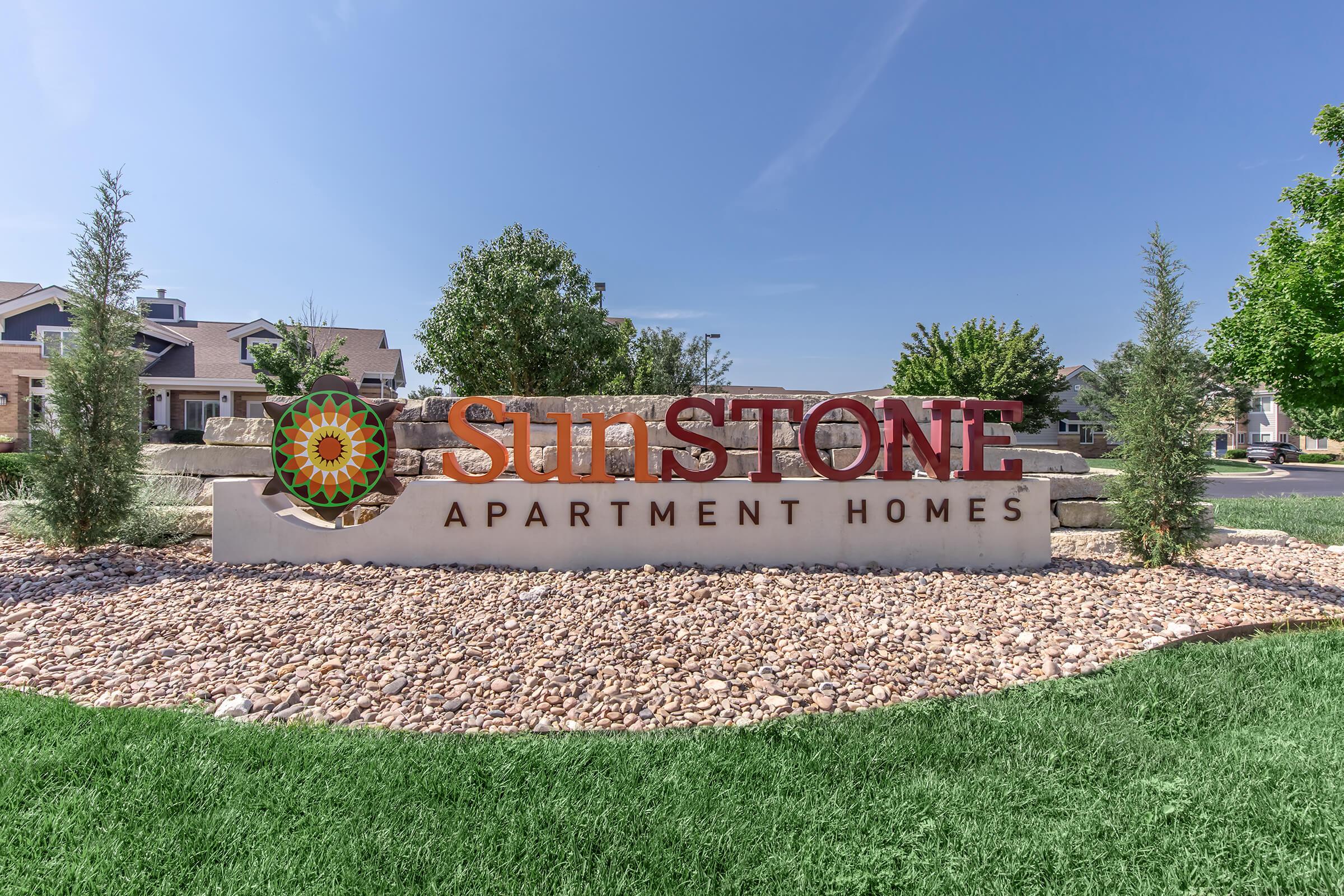 Sign for Sunstone Apartment Homes, featuring a colorful sun graphic, surrounded by decorative rocks and green grass. The background shows residential buildings under a clear blue sky.