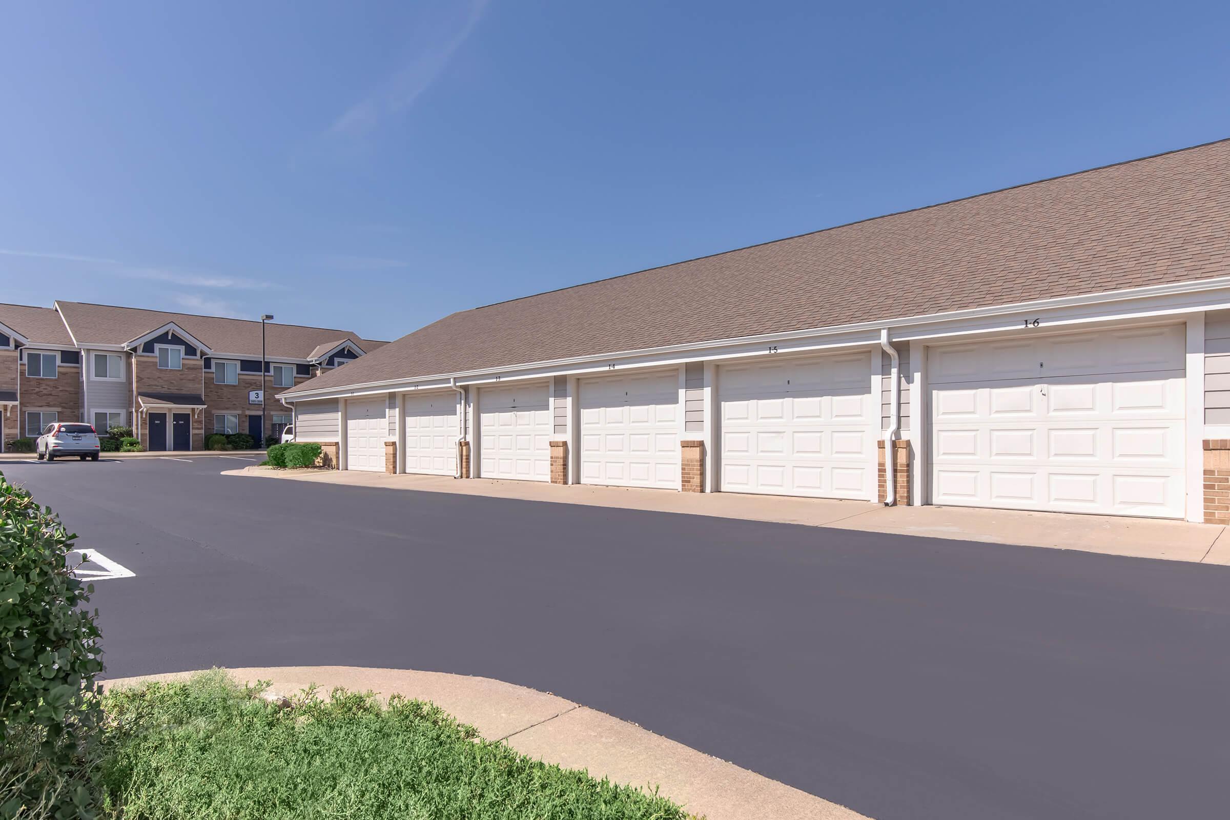 A row of white garage doors in a residential area, with a smooth asphalt driveway leading towards them. In the background, there are multi-story buildings with a brown roof and some greenery along the edges. The sky is clear and blue, indicating a sunny day.