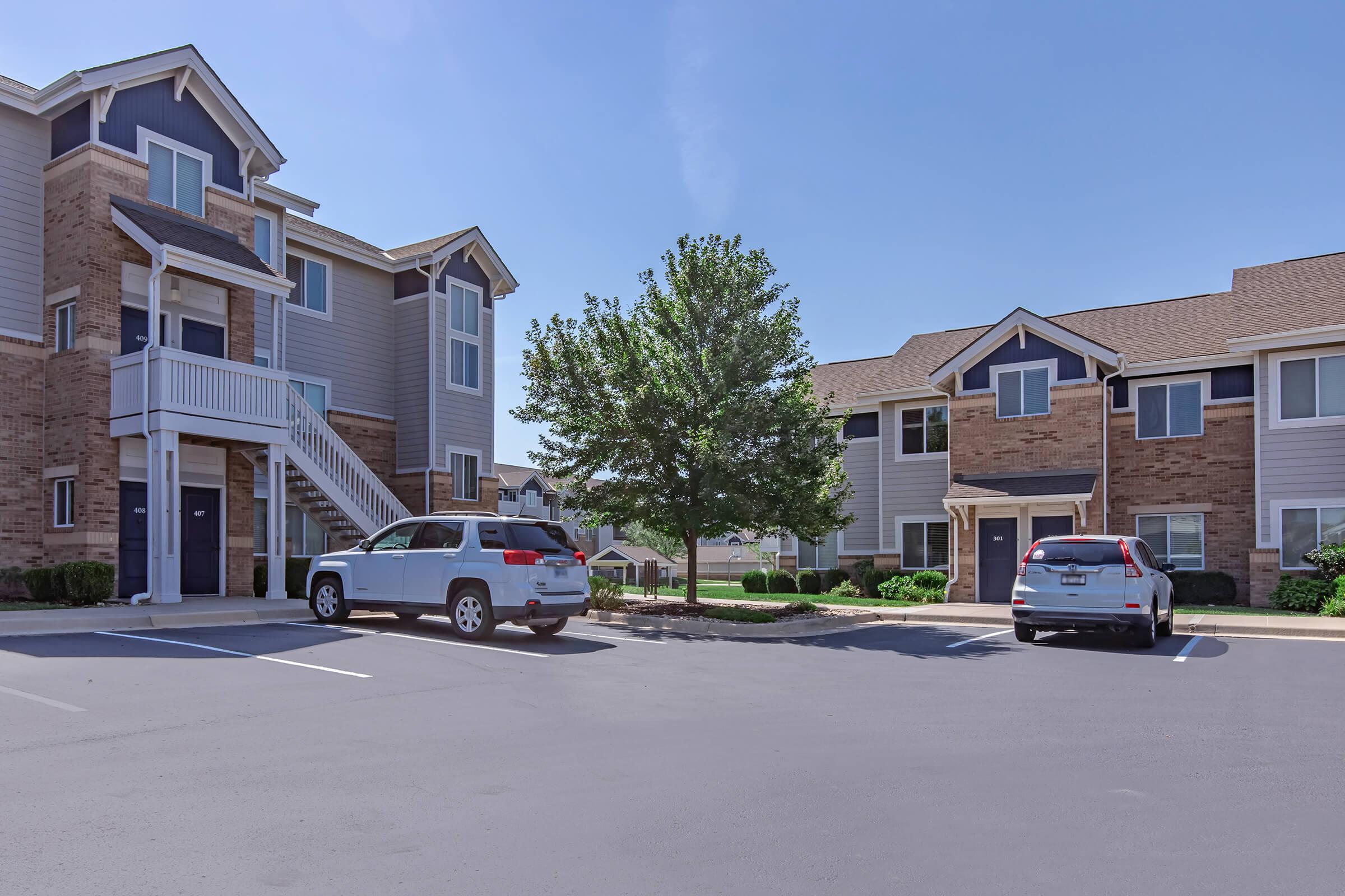 A residential apartment complex featuring two buildings, one with a staircase leading to the upper floor. The area includes a parking lot with parked vehicles and a tree, set against a clear blue sky.