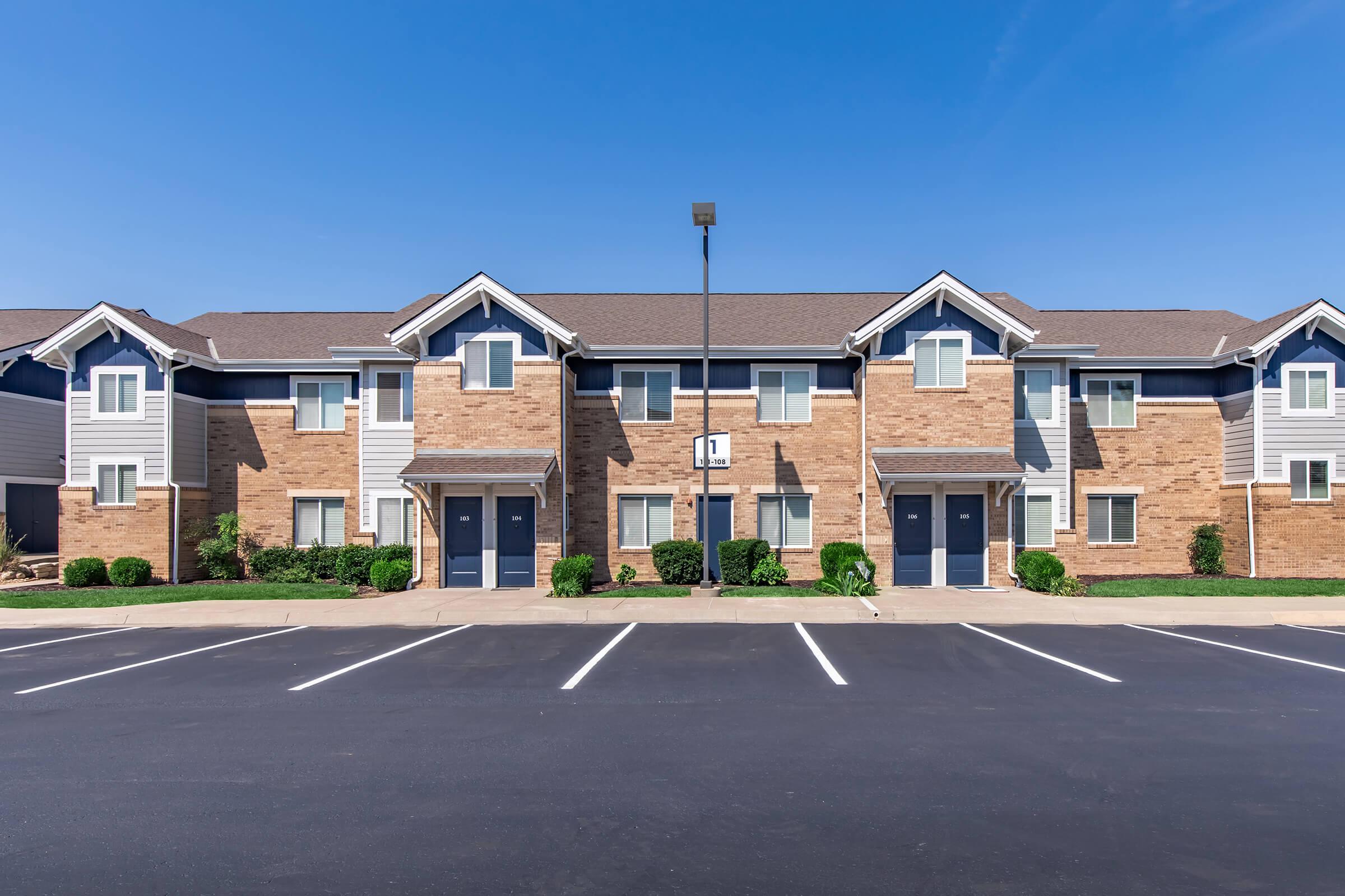 A row of modern brick apartment buildings with two stories. Each unit features blue front doors and symmetrical windows. The well-maintained landscape includes small bushes. The parking lot in front has marked spaces under a clear blue sky, indicating a bright and sunny day.