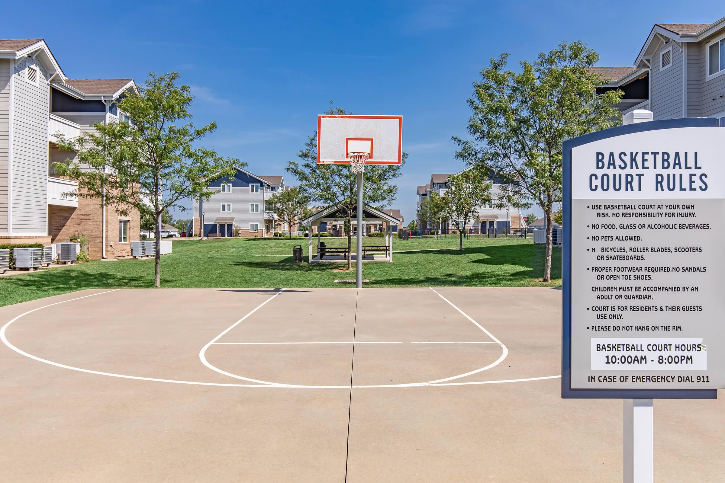 A basketball court with rules signage is prominently displayed. The court is surrounded by green grass and residential buildings in the background. The sign lists basketball court rules and hours, with an emergency contact number provided. Bright blue sky overhead complements the outdoor setting.