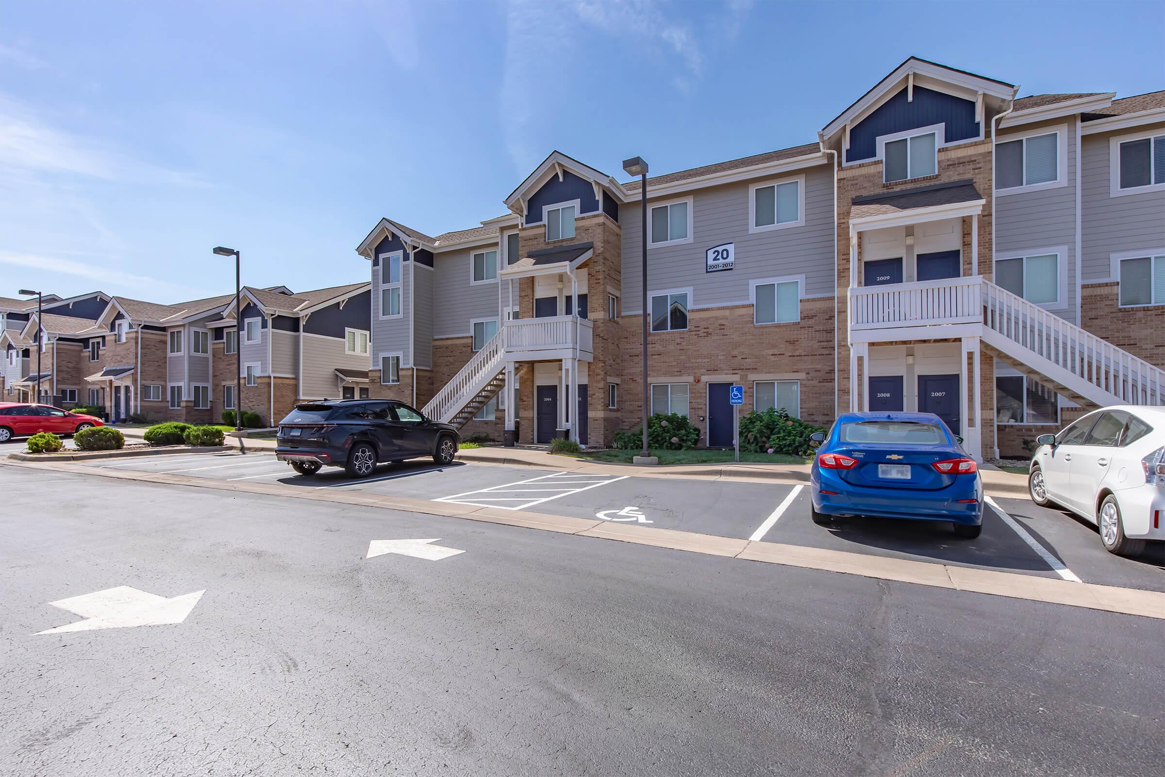 A multi-unit residential building with a light-colored exterior, featuring several staircases. The parking lot includes a few parked cars and designated spaces, with clear blue skies overhead. The building is surrounded by well-maintained landscaping.