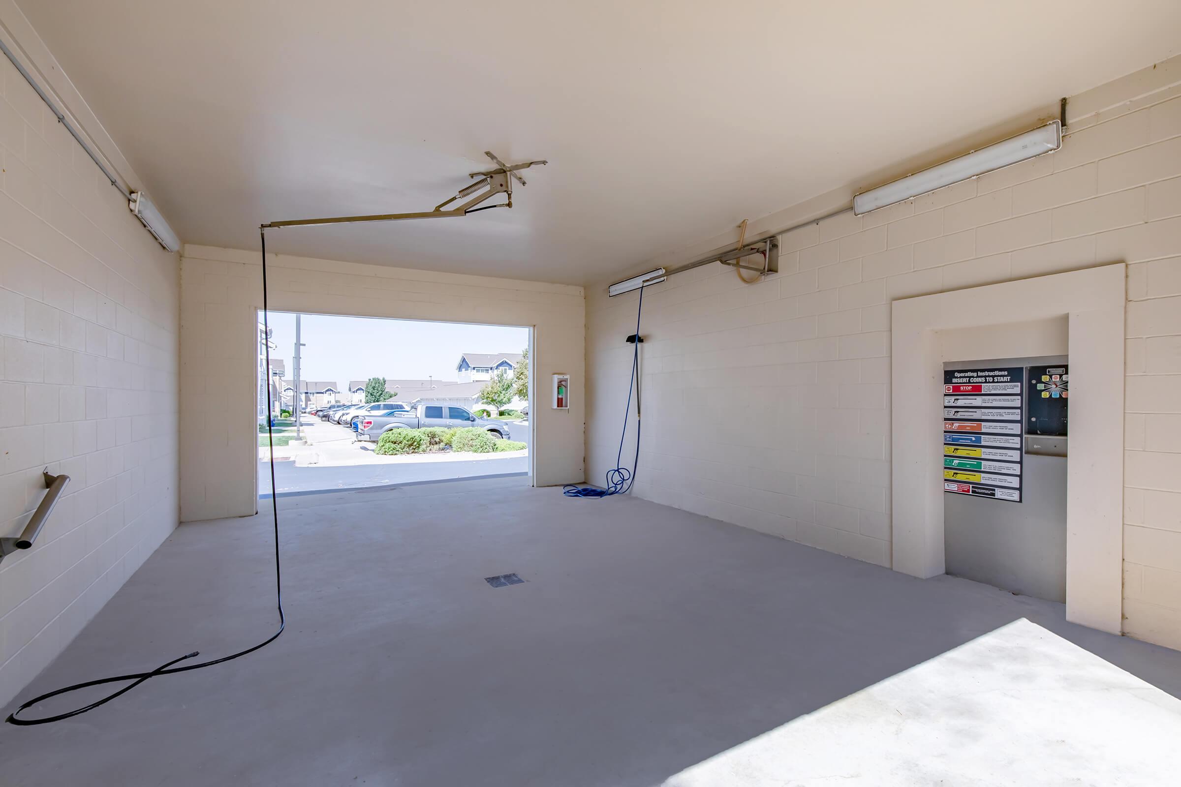 An empty car wash bay featuring a large open entrance with a view of parked cars outside. There are hoses and cleaning equipment hanging from the ceiling, and a payment station with various car wash options is mounted on the wall. The interior is well-lit and has a plain, minimalist design.