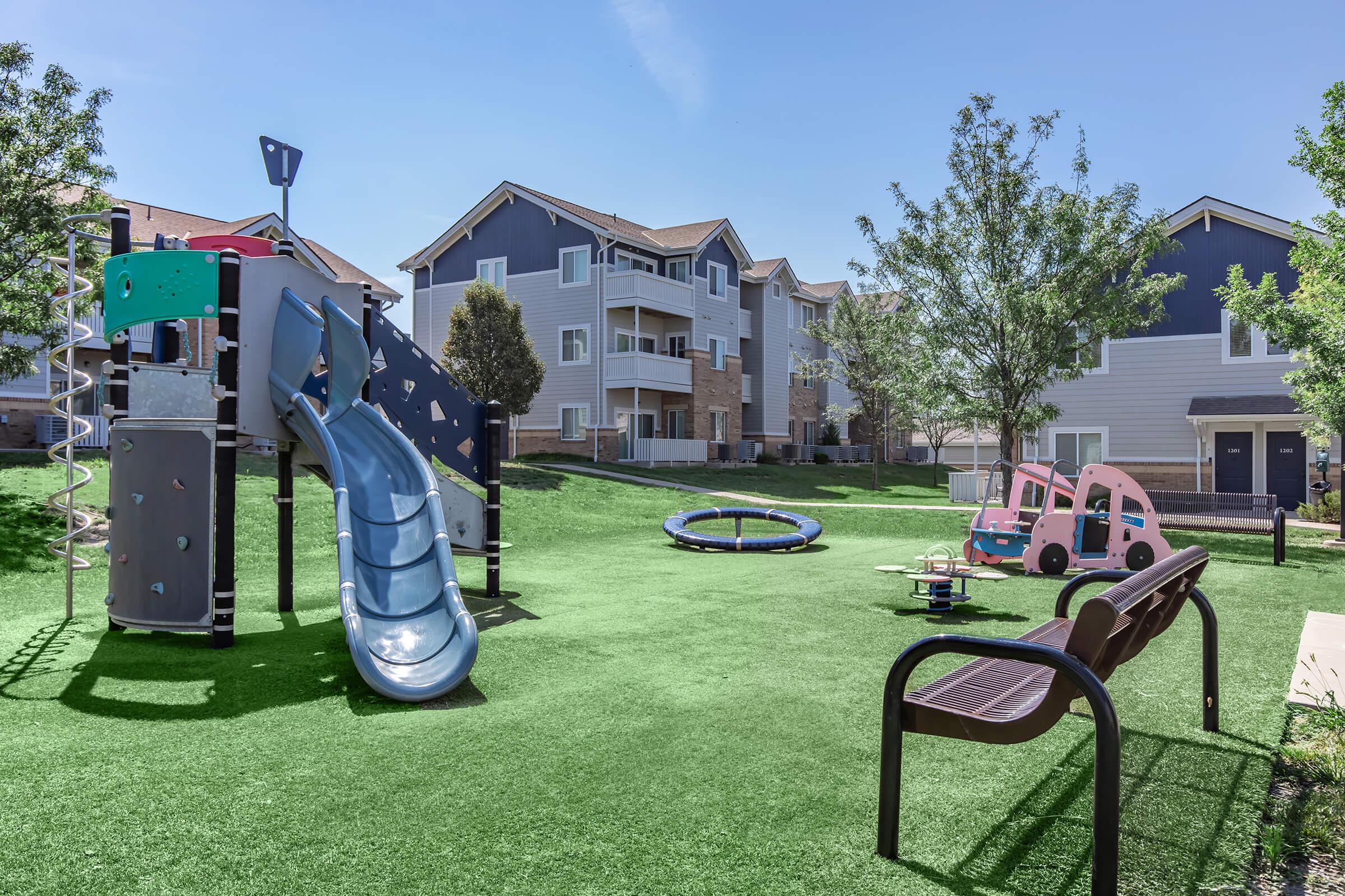 A colorful playground featuring a slide, climbing structure, and a trampoline, set on green turf. Nearby, there are benches and toy cars, with modern apartment buildings in the background under a clear blue sky.