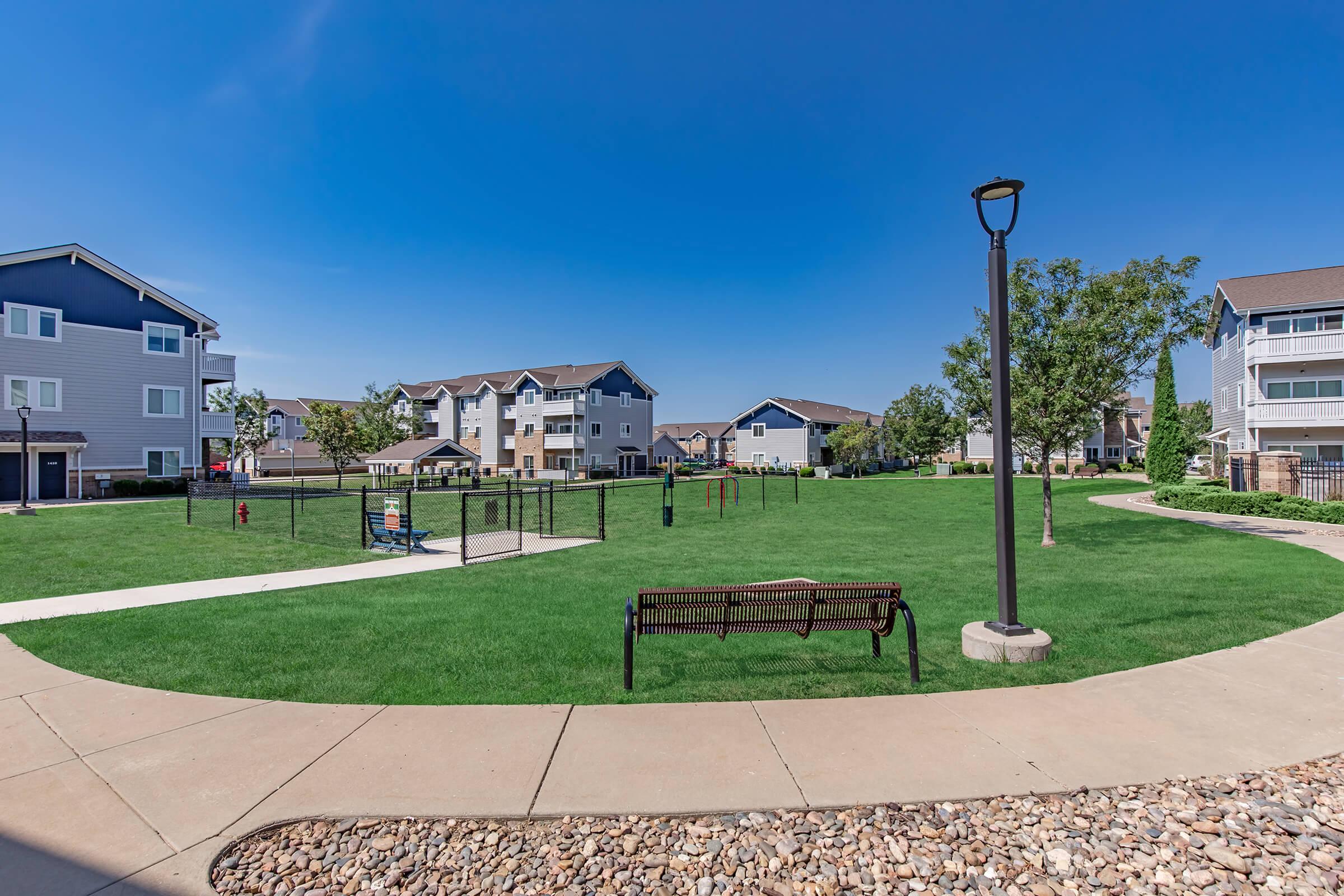 A wide view of a green park area featuring a walking path, a bench, and a playground nearby. In the background, several multi-story residential buildings are visible under a clear blue sky, surrounded by trees and landscaped grass.