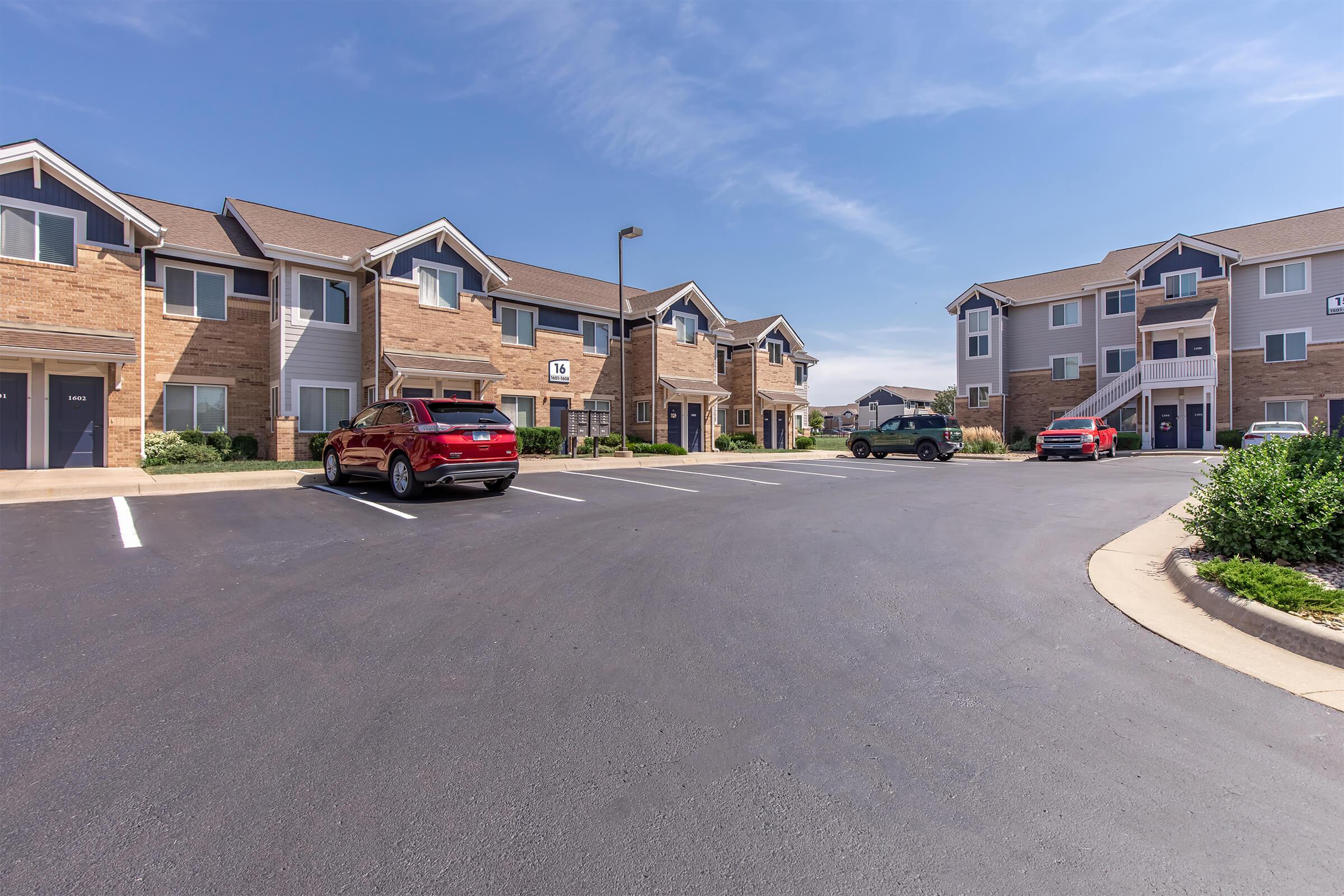 A residential apartment complex featuring two-story buildings, with several parked cars in the foreground. The parking area is paved and surrounded by green shrubs. The sky is clear with a few clouds, indicating a bright day.