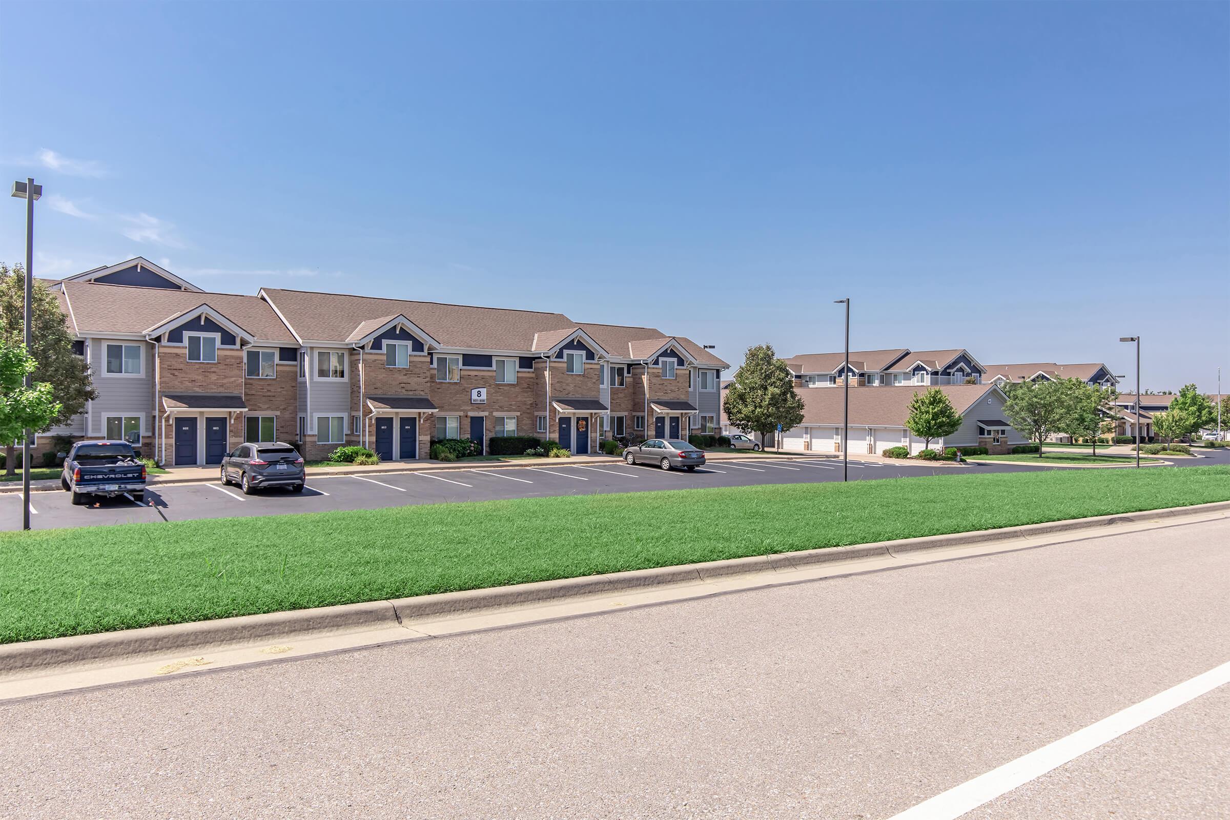 A street view of a residential area featuring two buildings with multiple apartment units. The buildings have a combination of brick and siding exteriors, with parking spaces in front. A well-maintained grassy area is visible in the foreground, and a clear blue sky completes the scene.
