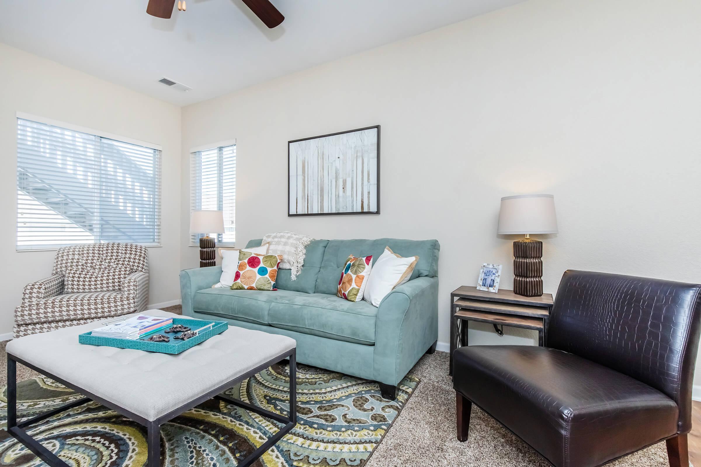 A cozy living room featuring a light blue sofa with colorful pillows, a patterned area rug, and a decorative coffee table. There is an armchair with a checkered pattern, a side table with a lamp, and artwork on the wall. Natural light filters through blinds, creating a warm atmosphere.