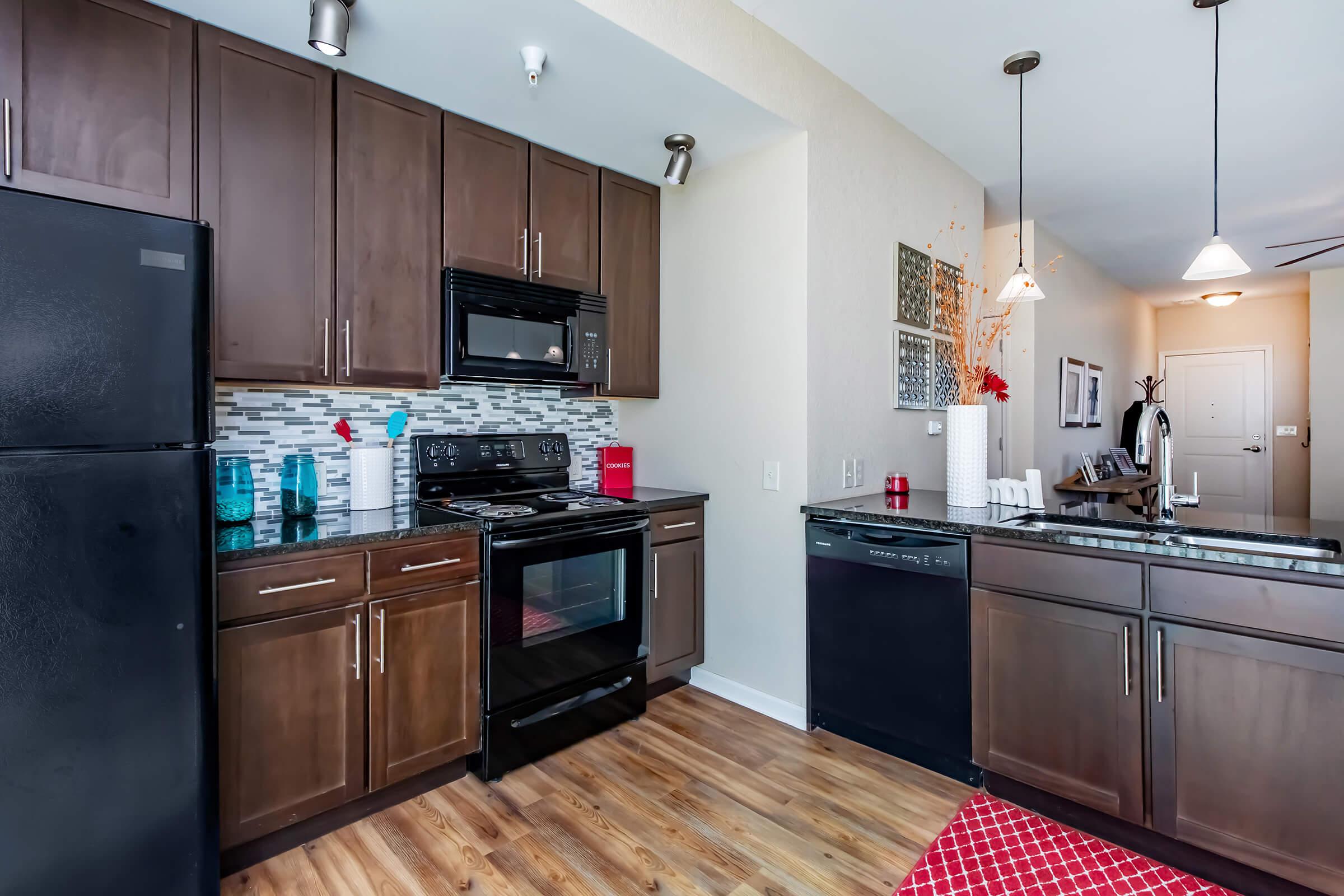 A modern kitchen featuring dark wooden cabinets, a black refrigerator, an oven, and a dishwasher. The backsplash is made of small tiles, and there are decorative items on the counter. A red area rug lays on the wooden floor, and pendant lights hang from the ceiling, adding warm lighting to the space.