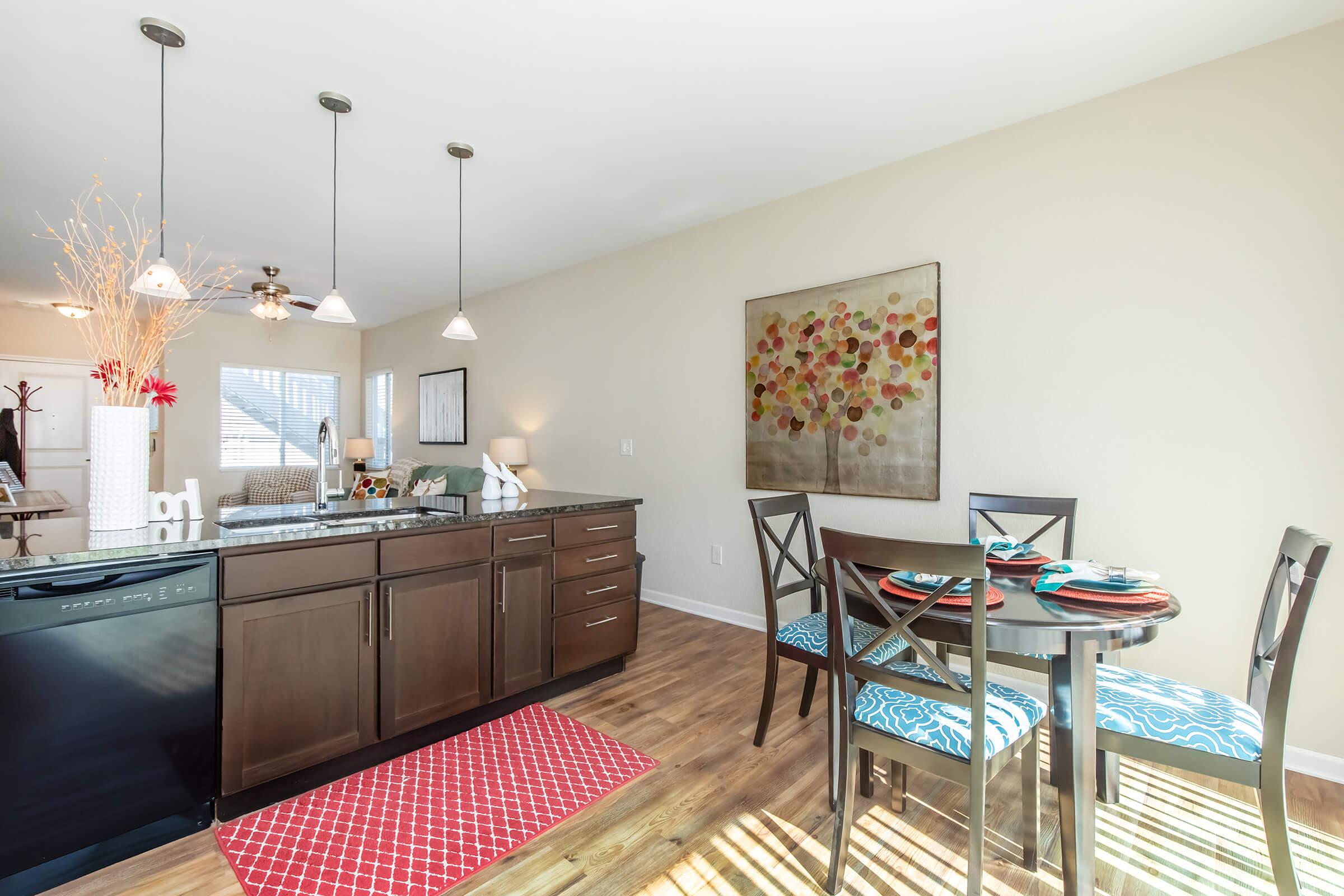 Modern kitchen and dining area featuring a dark wood cabinetry, a black dishwasher, and a round table with colorful place settings. The floor is wooden, and there's a red area rug. Bright pendant lights illuminate the space, complemented by a painting with colorful circular patterns on the wall.