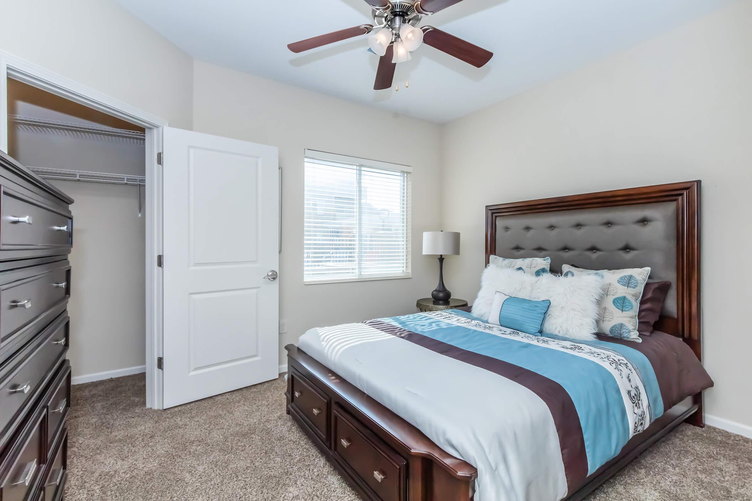 A neatly arranged bedroom featuring a large bed with a light blue and brown bedding, decorative pillows, and a dark wooden headboard. There is a dresser, a bedside lamp, and a closet visible. The walls are painted light colors, and a ceiling fan hangs above. Natural light enters through a window.